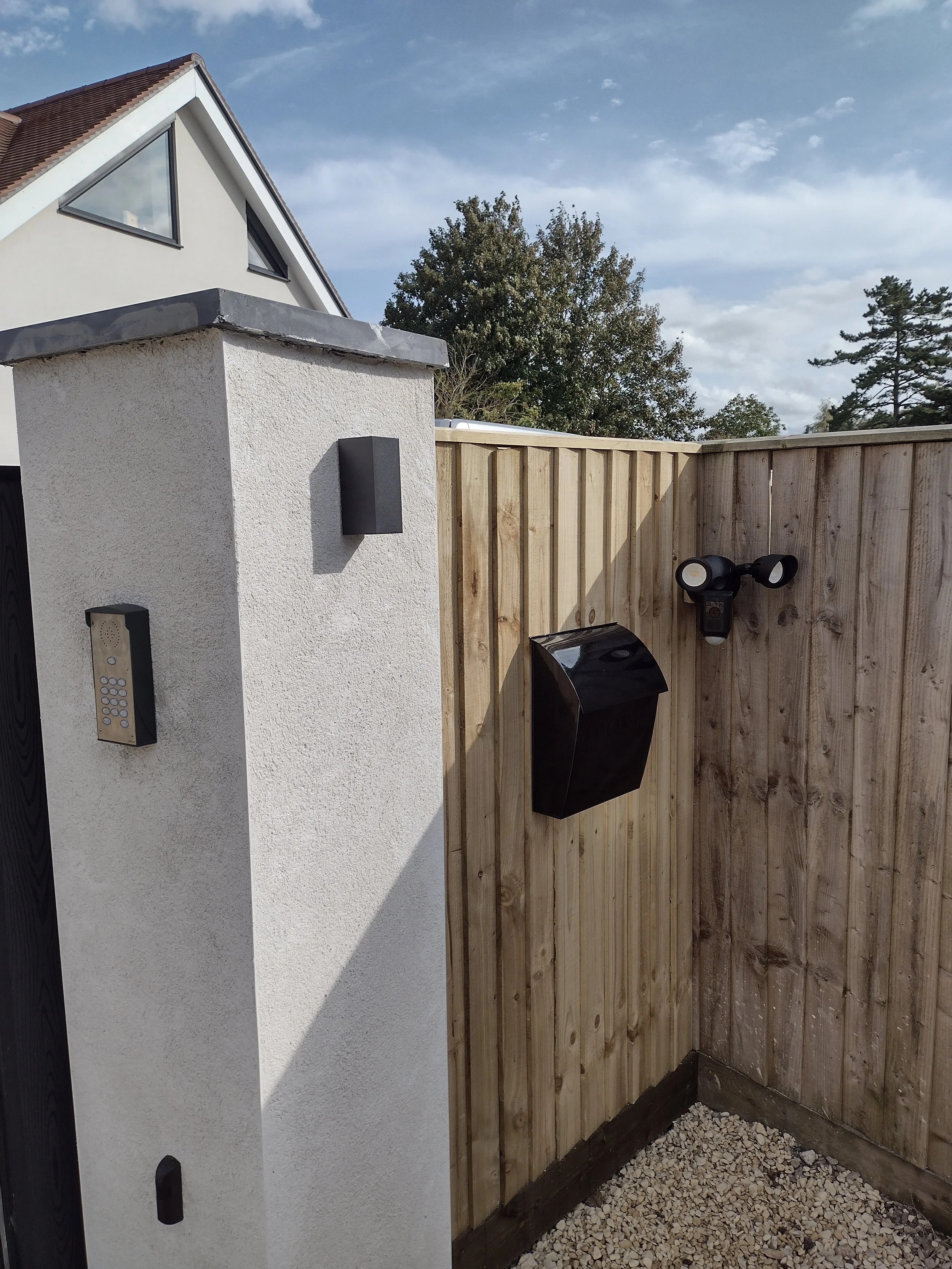 An outdoor corner with a wooden fence, a white textured wall, a black trash bin, and outdoor lights mounted on the fence. The house has a sloped roof with windows, and the sky is partly cloudy with trees in the background.