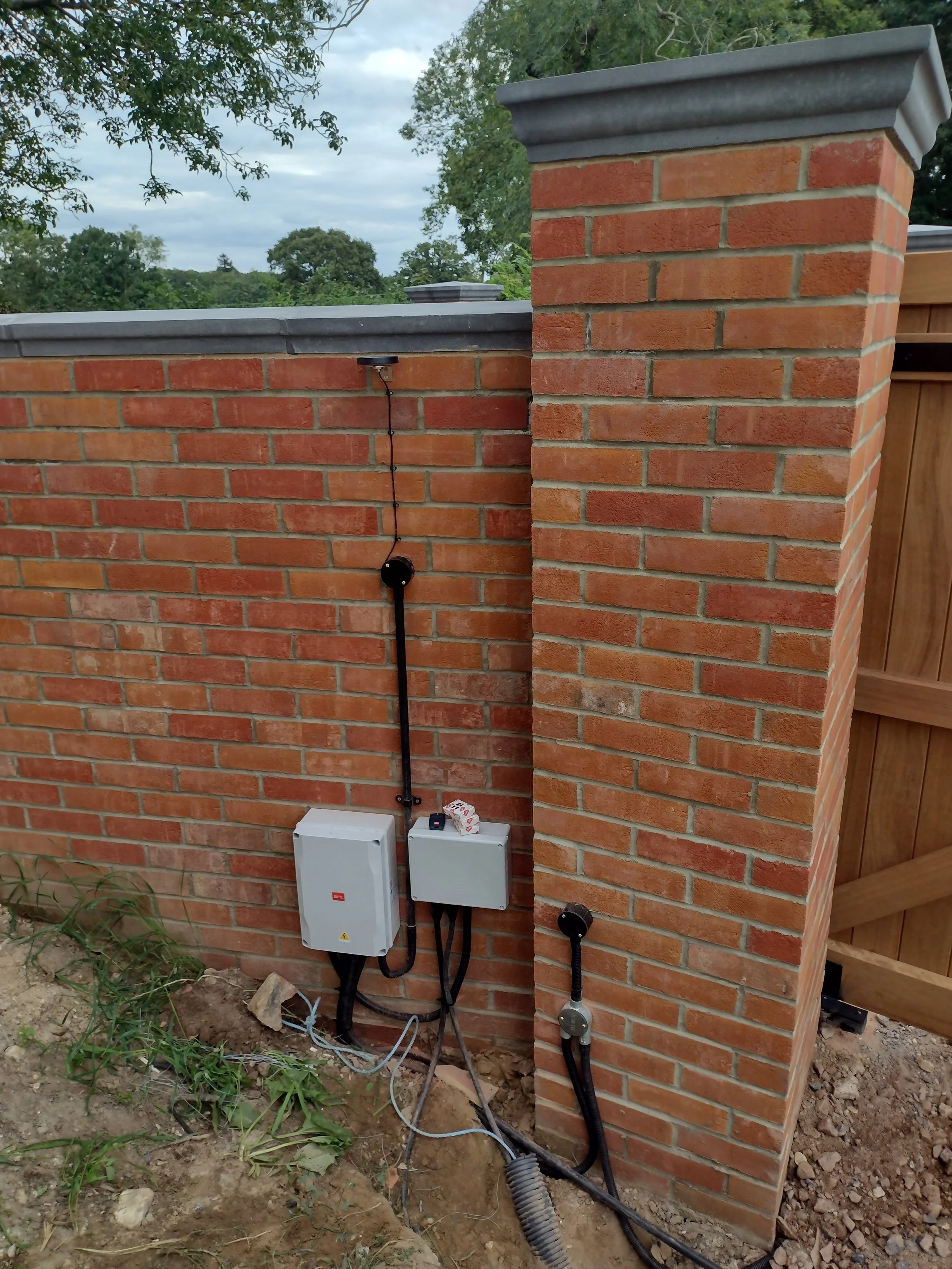 A brick wall with an electrical panel, wires, and a conduit on the ground.