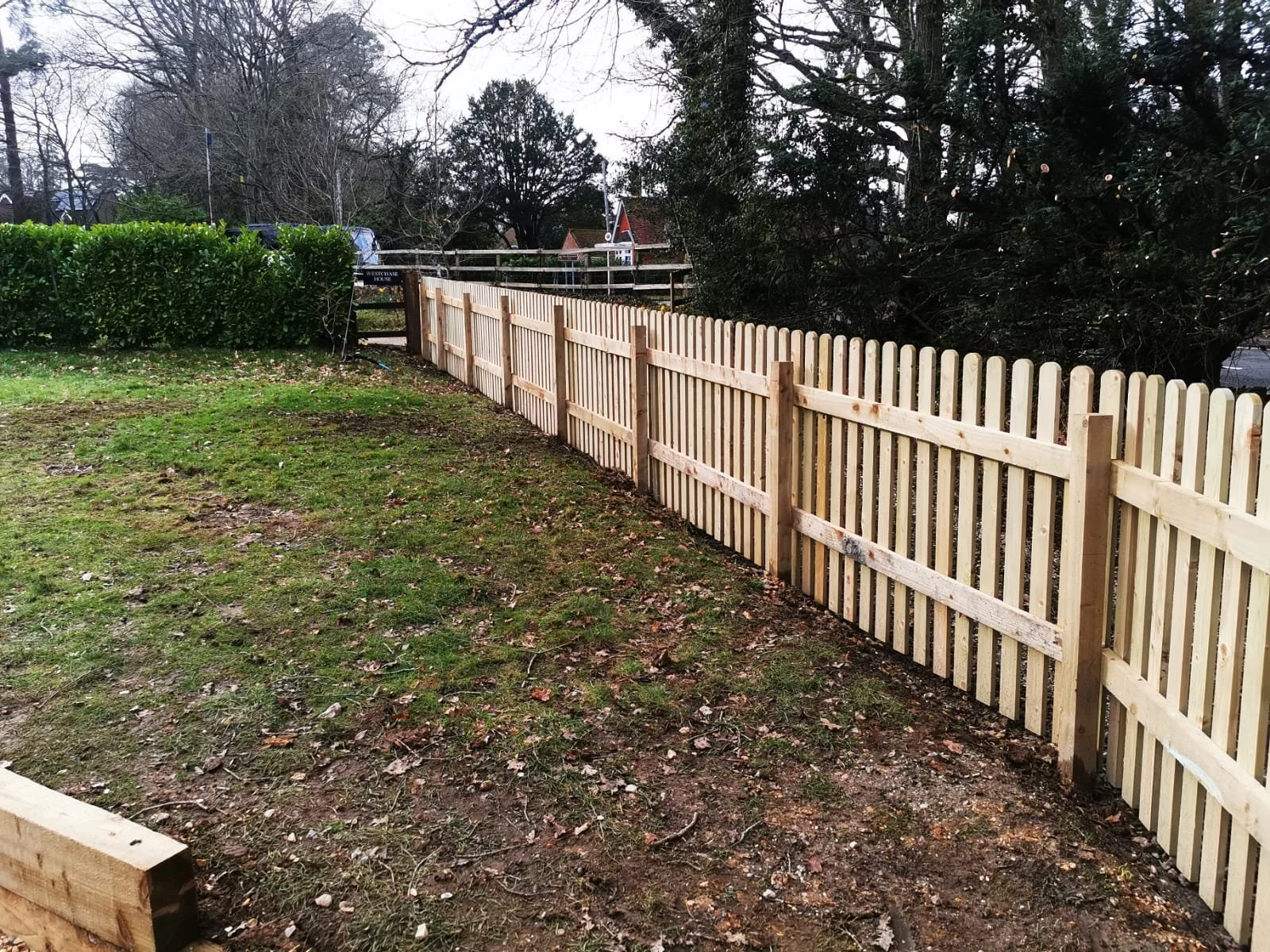 A wooden picket fence being installed in a backyard with grass, trees, and a hedge line in the background.