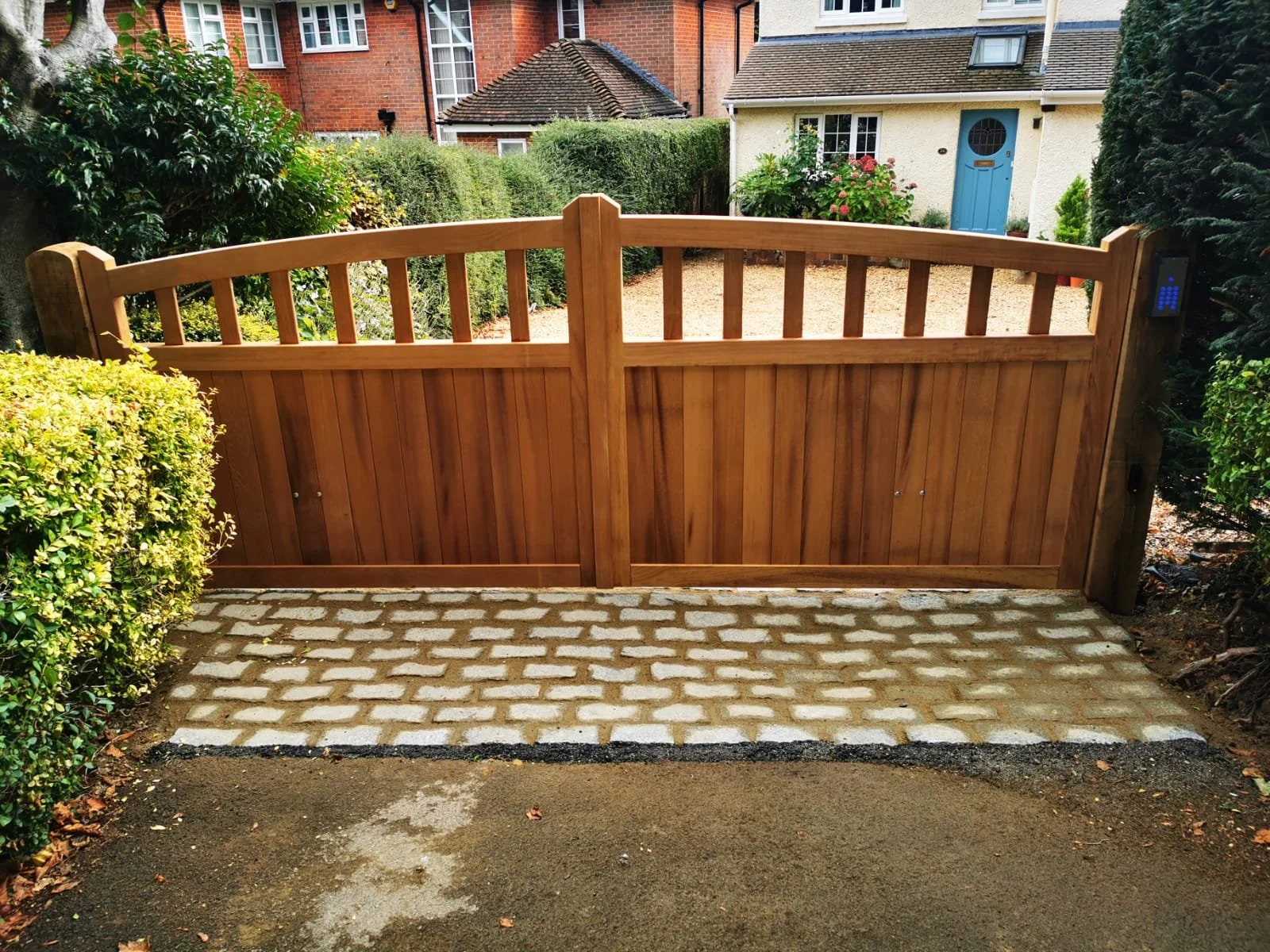 Wooden driveway gate with an electronic keypad entry system on the right post, set in front of a gravel driveway and a beige house with a blue door, surrounded by greenery and a brick-paved walkway.