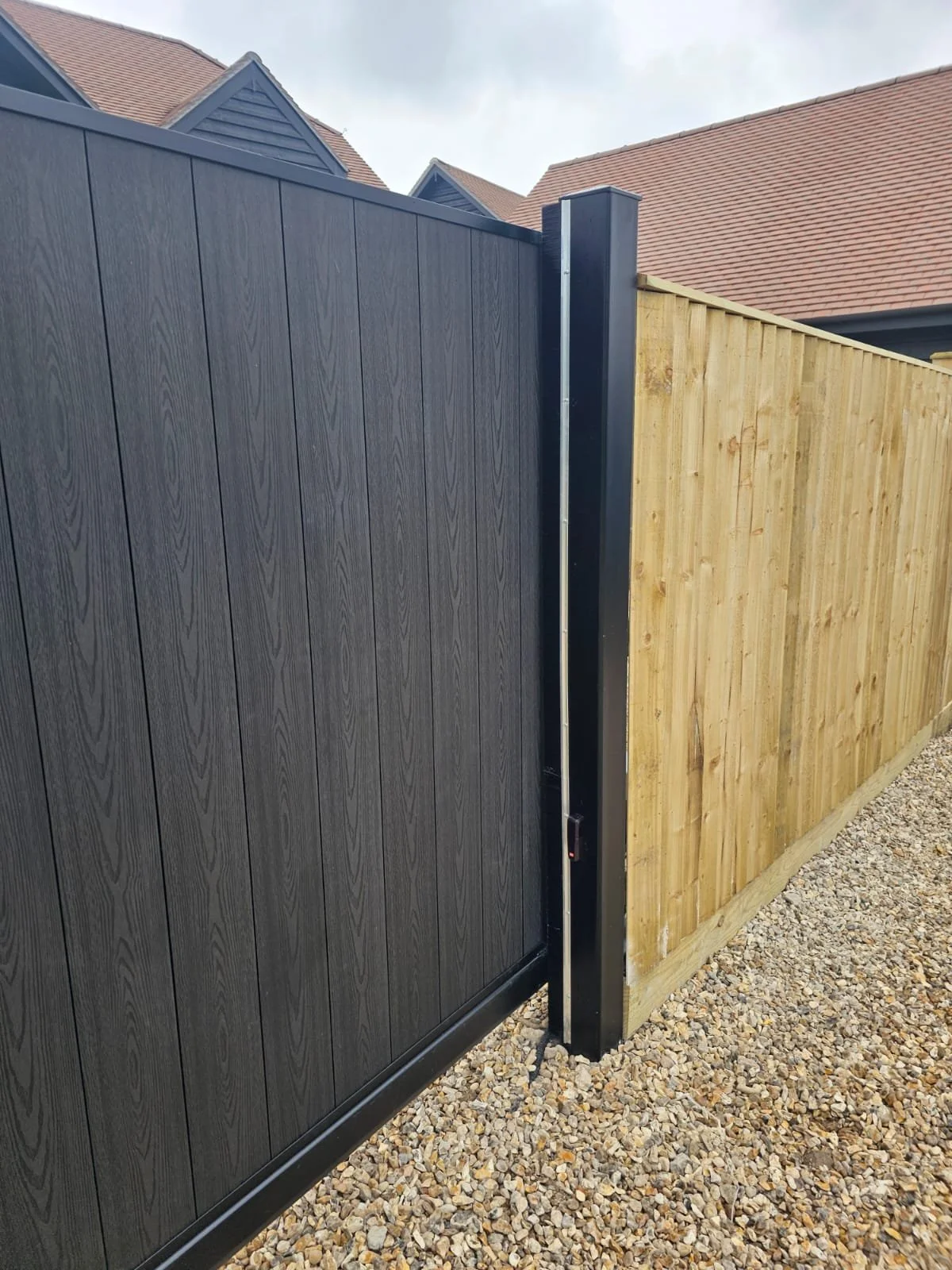 A black sliding gate next to a wooden fence, with a gravel ground and house roofs in the background.