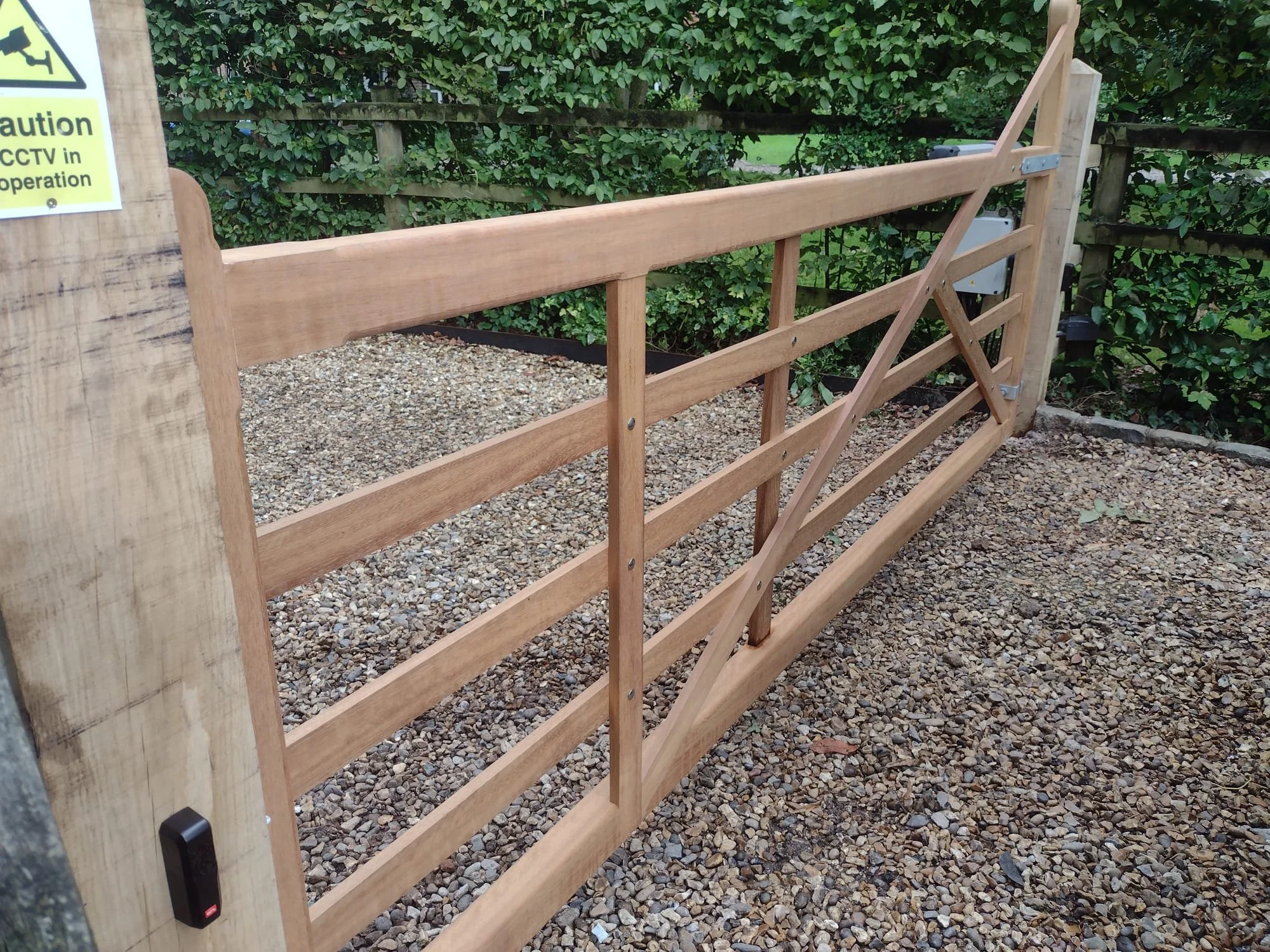 Wooden gate with horizontal slats, secured with metal brackets, near a gravel ground and green foliage in the background.