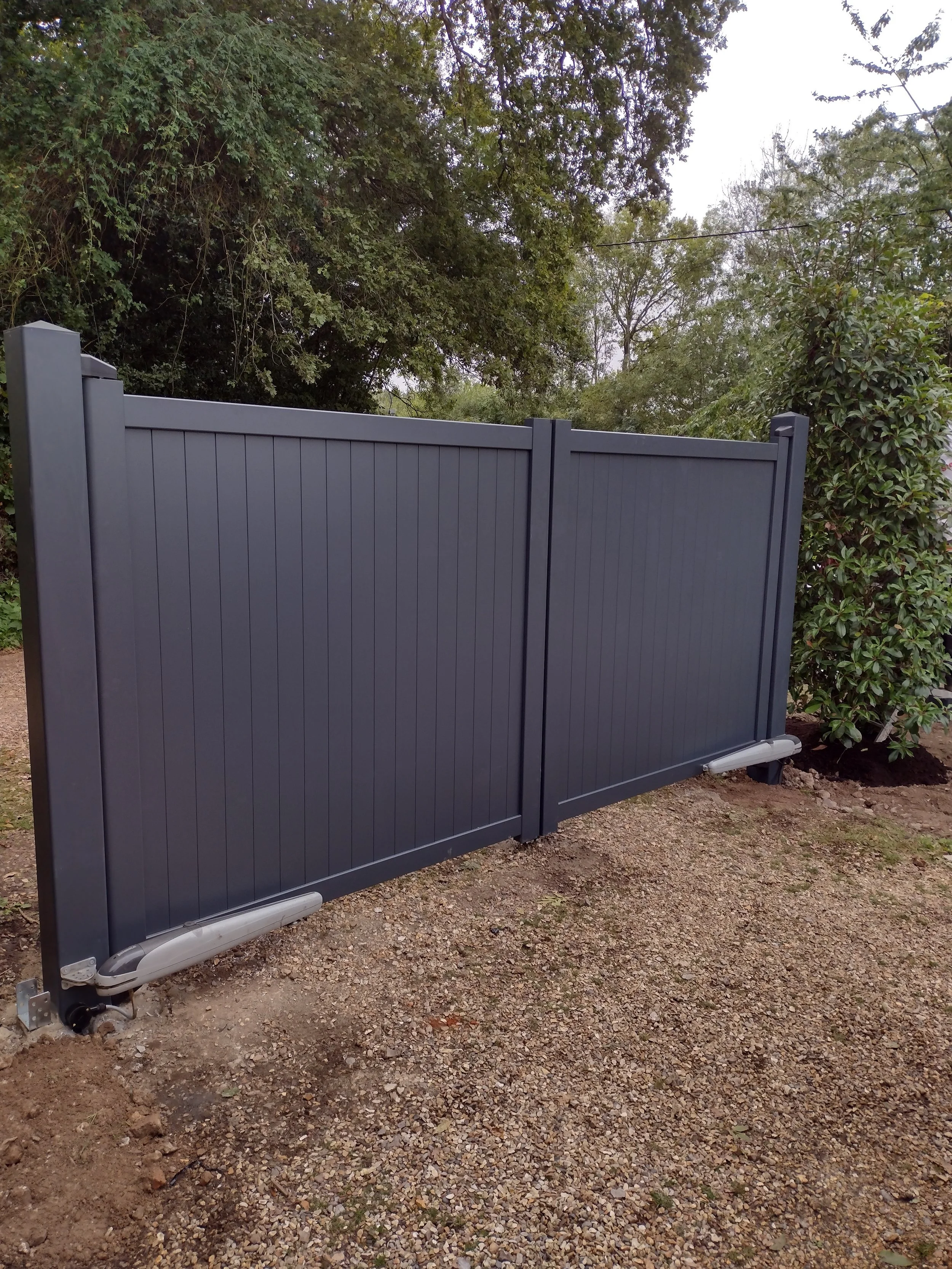 A gray, metal sliding gate installed in an outdoor area with gravel ground and surrounded by green trees and bushes.