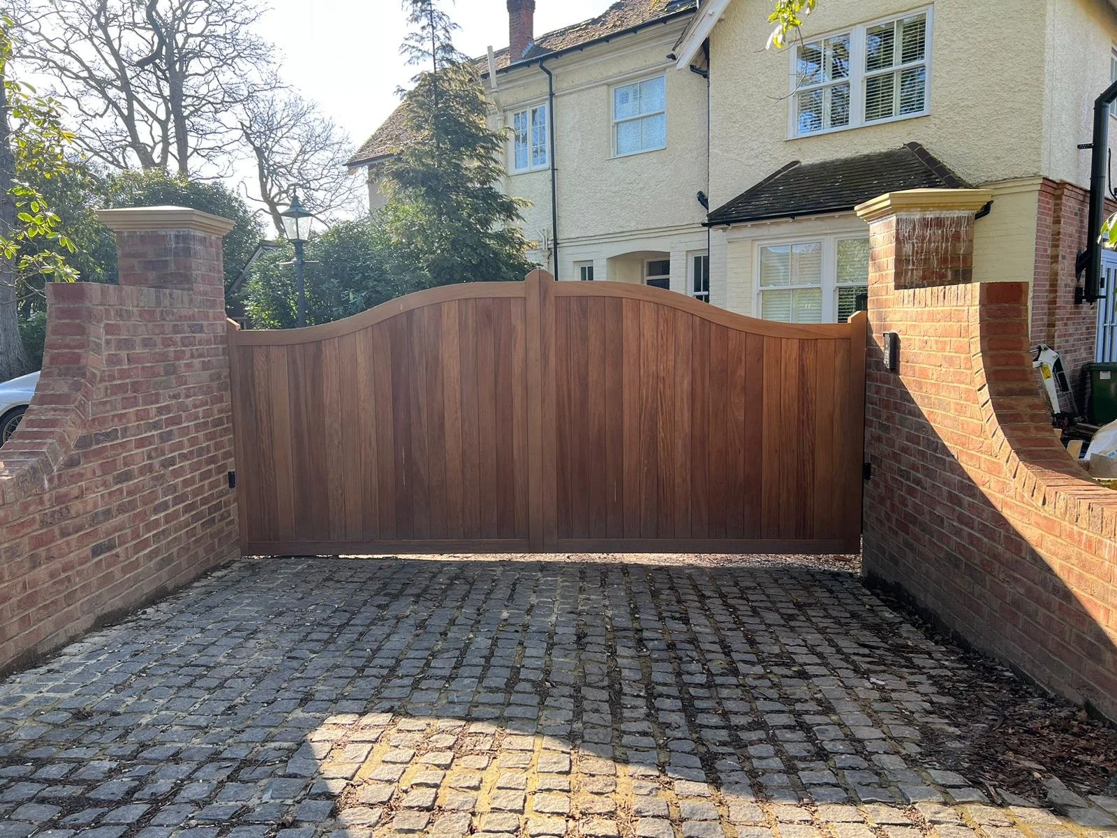 A wooden gate with brick pillars on either side, situated in front of a house with yellow walls and multiple windows.
