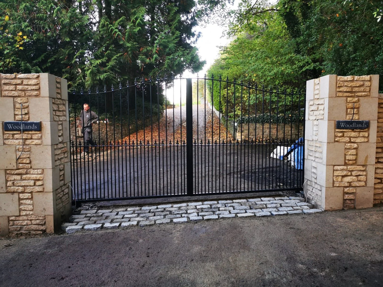 A black wrought iron gate with stone pillars on each side, marked with 'Woodlands', blocking access to a pathway lined with green trees.