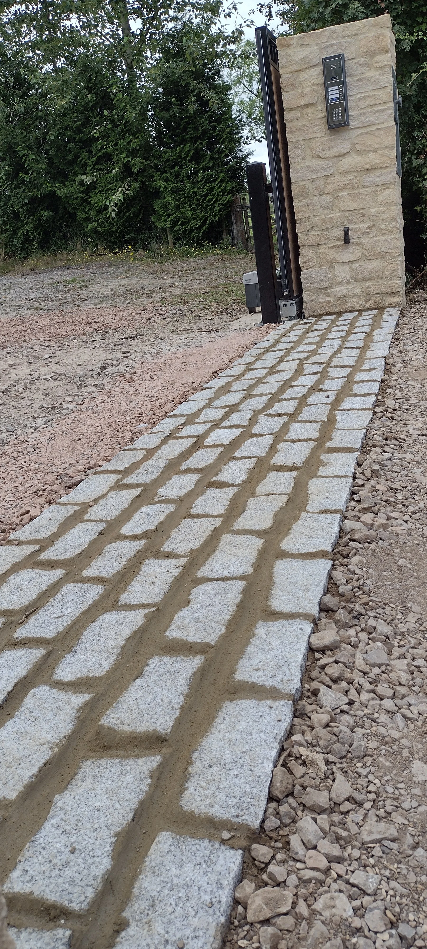Pathway made of bricks with a concrete border leading to a stone wall entrance equipped with an electronic doorbell and keypad, surrounded by gravel and greenery.