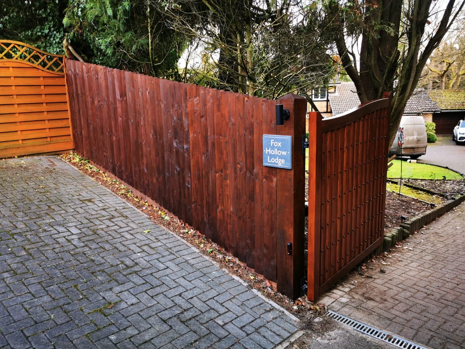 A wooden fence with a sign that reads 'Fox Hollow Lodge.' The fence surrounds a property with a large tree, and there are cars parked on the street in the background.