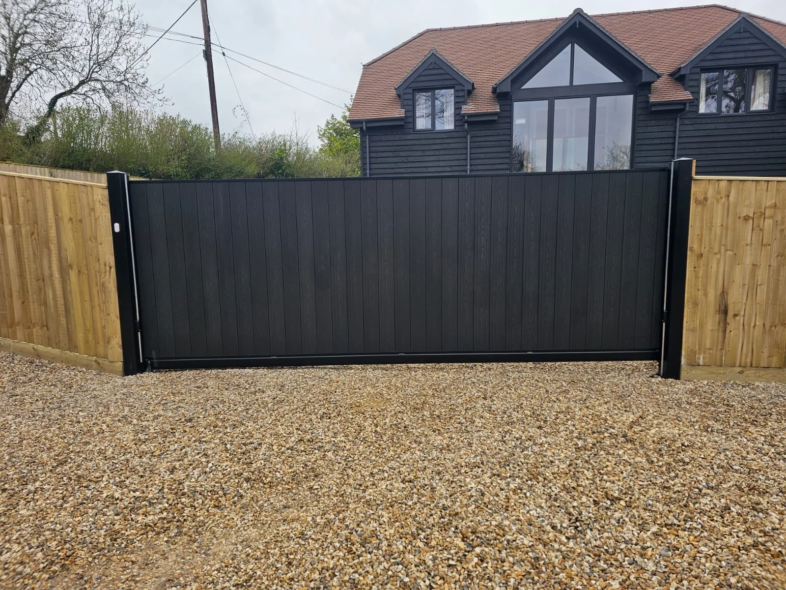 A black metal gate with a wooden frame on each side, situated on a gravel driveway in front of a two-story house with black siding and large windows.