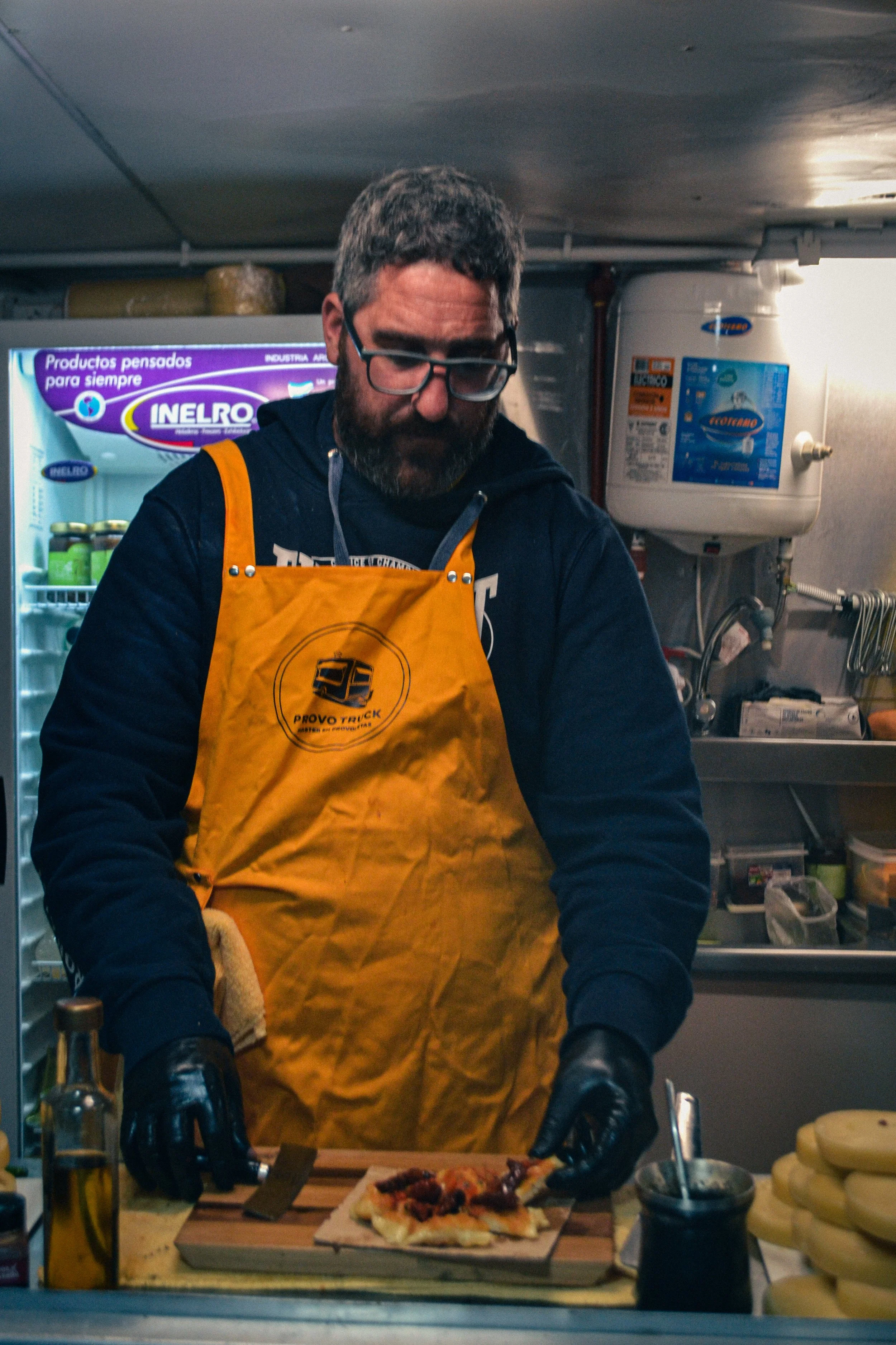 Un hombre con barba y lentes, vestido con un delantal naranja, prepara una pizza en una cocina.