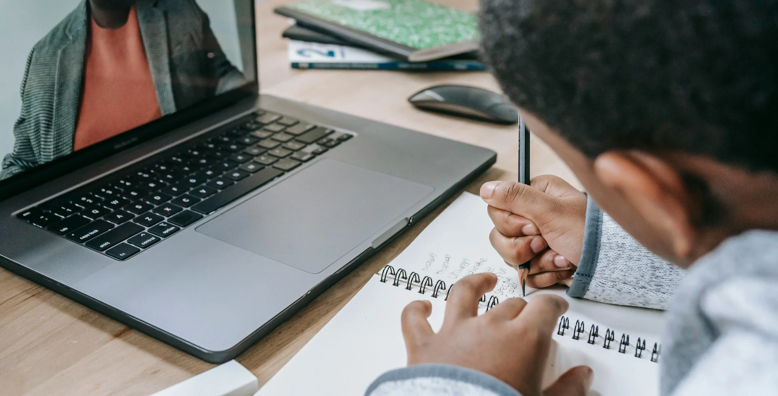 Person taking notes with a pen on a notepad while sitting at a desk with a laptop, mouse, and notebooks.