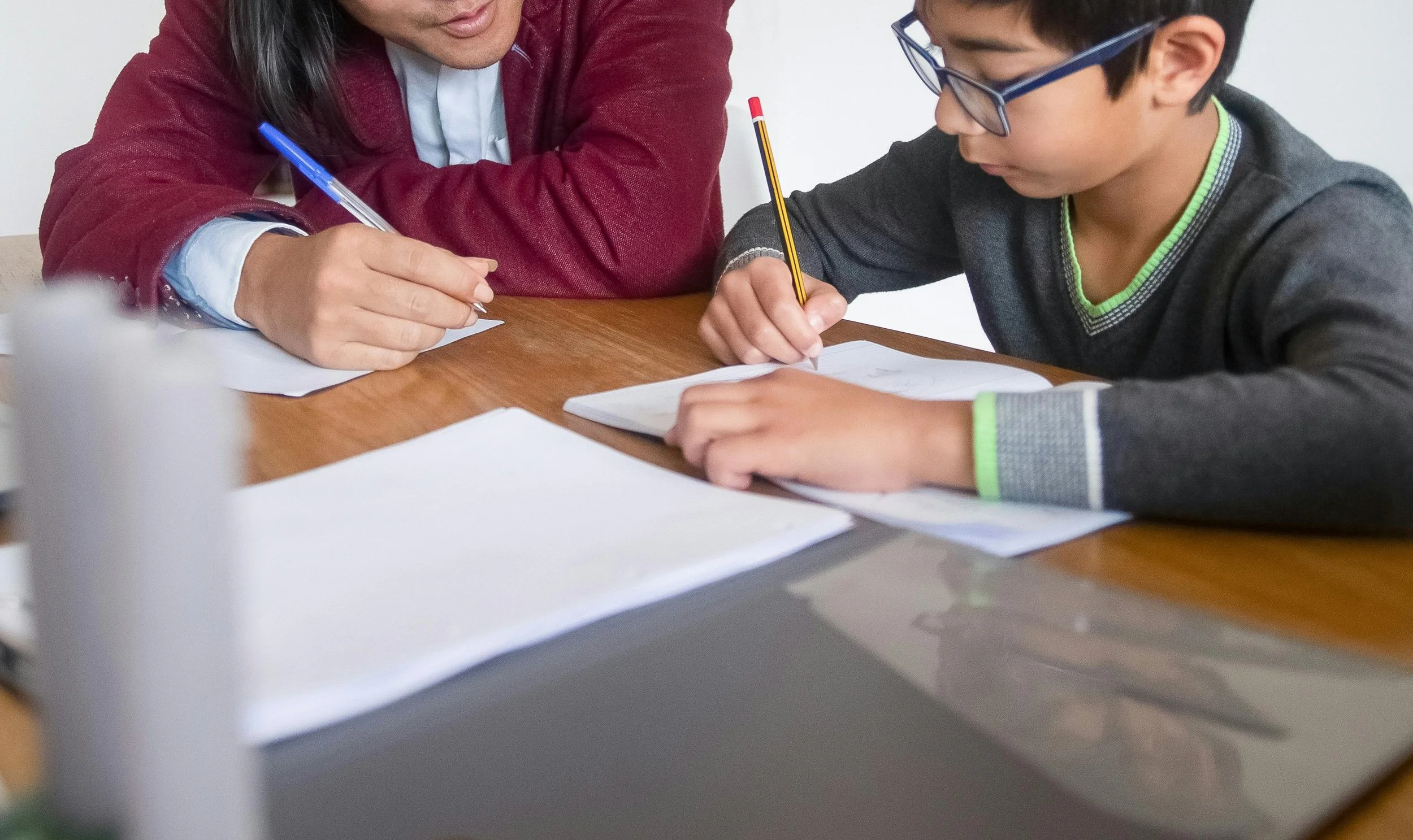 A woman and a young boy sitting at a wooden table, engaging in writing or drawing activities, with papers and notebooks in front of them.