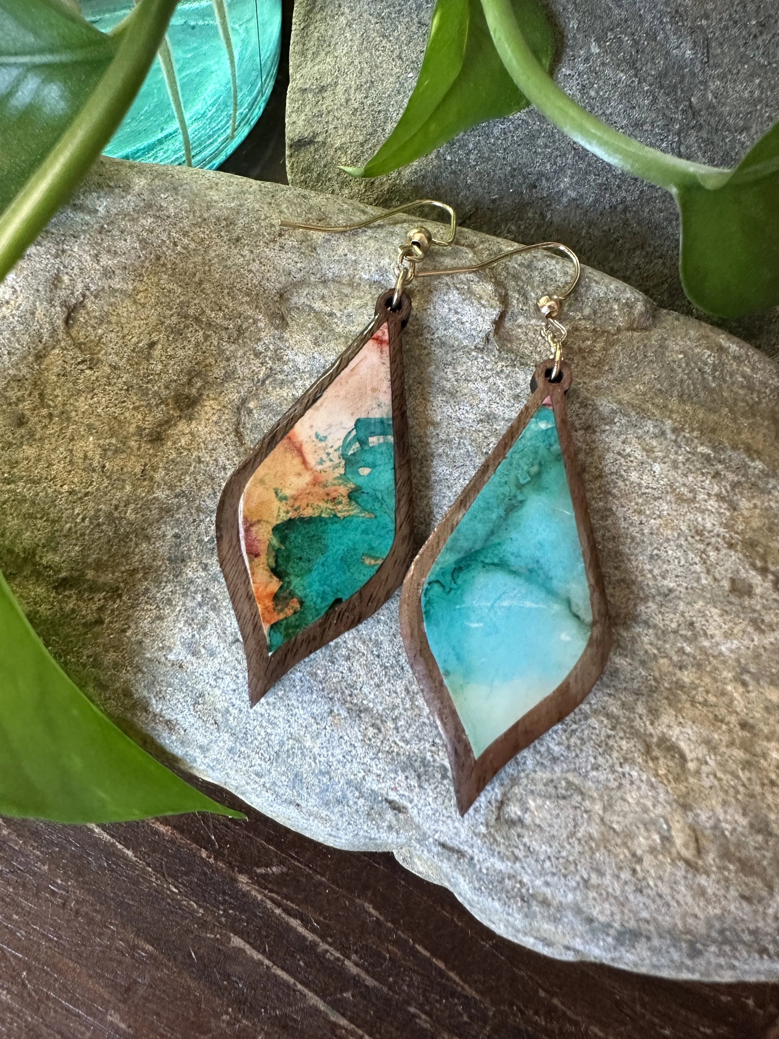 Colorful teardrop-shaped earrings with abstract watercolor design in green, blue, orange, and pink, resting on a textured stone surface surrounded by green leaves.