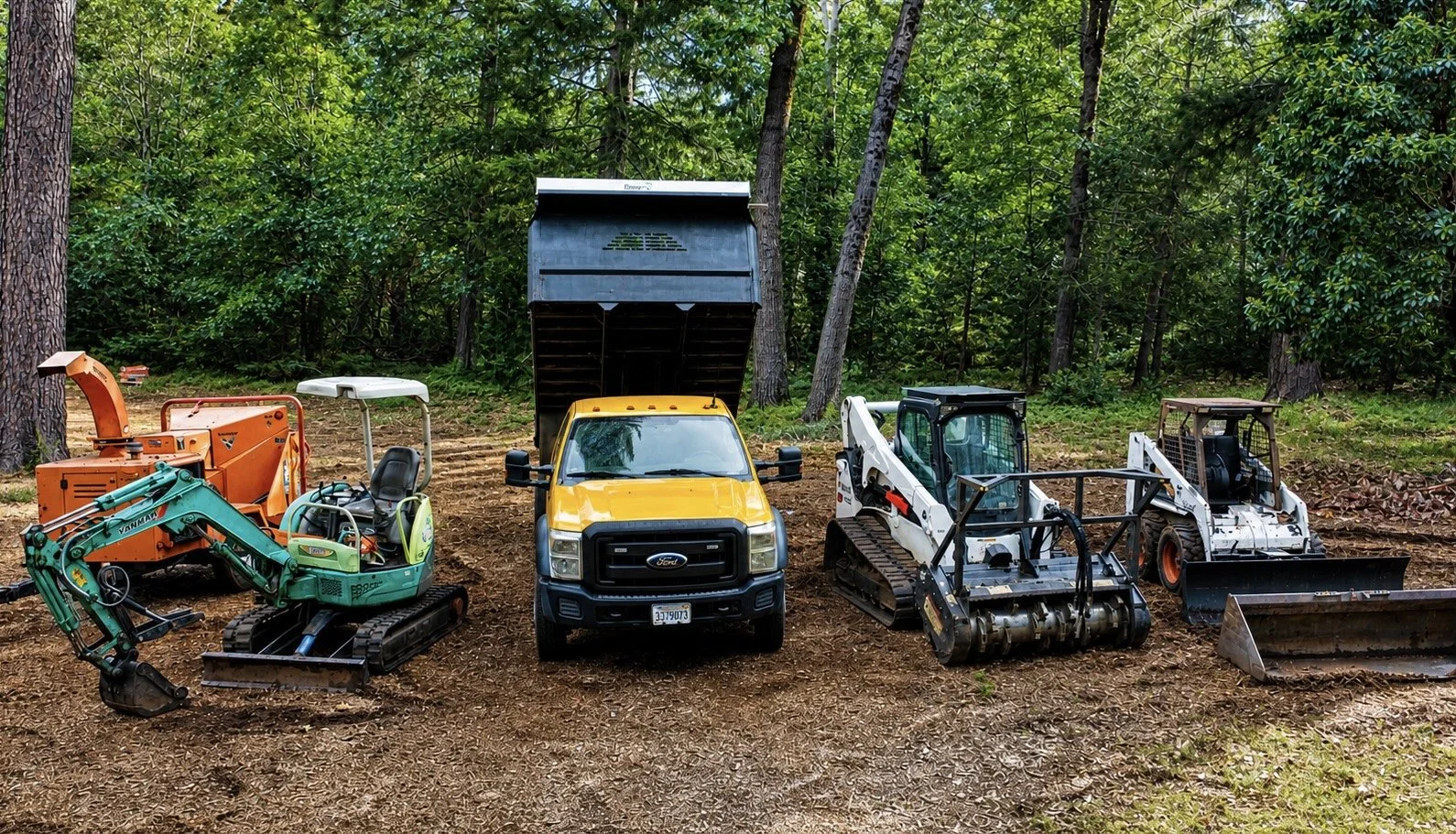 Array of construction and landscaping equipment on dirt in a wooded area, including a green mini excavator, a yellow and black Ford truck, an orange woodchipper, a white skid-steer loader, and a white compact loader.