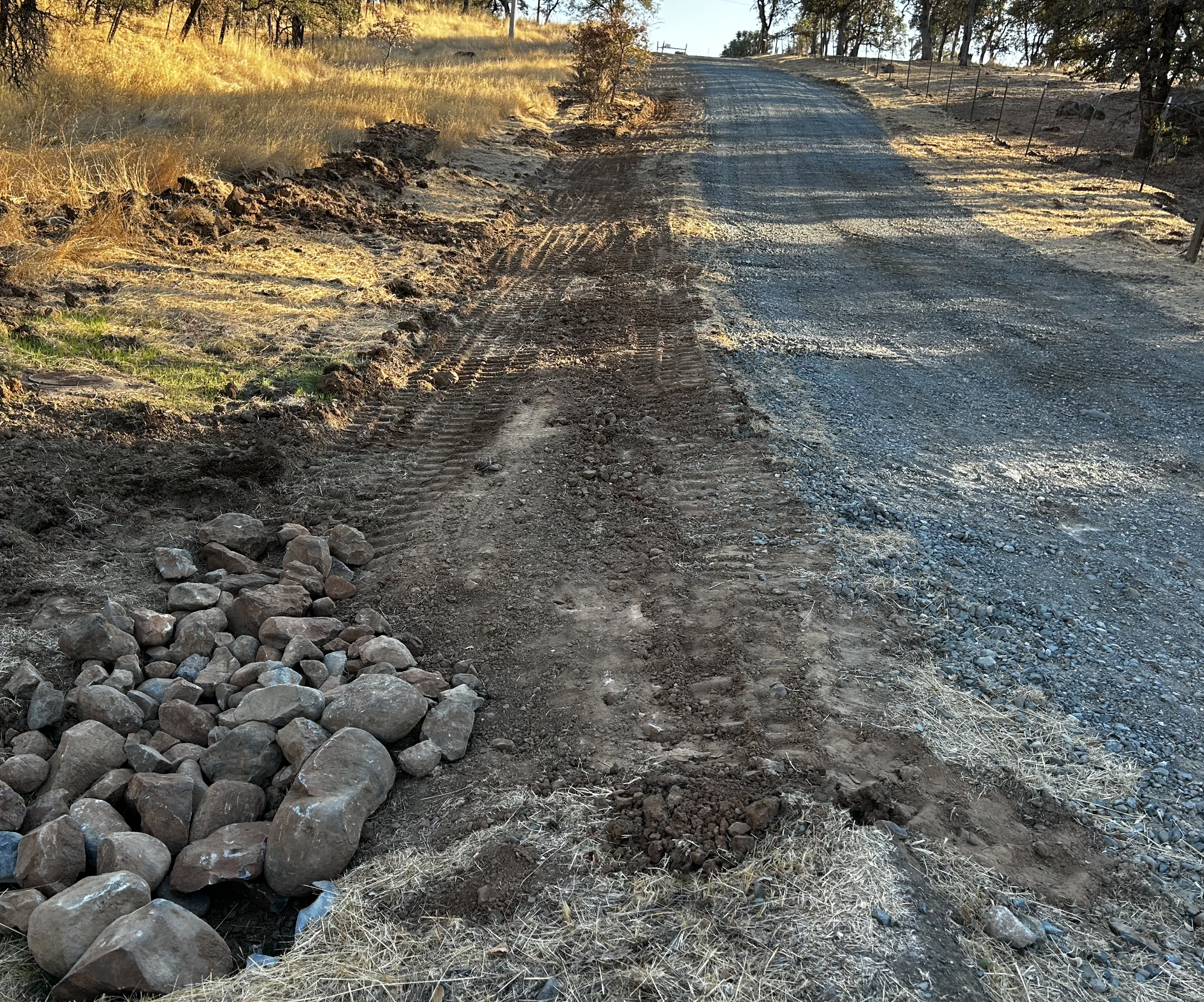 A dirt pathway with tire tracks, bordered by rocks on the left and fenced area on the right, on a hill with trees and dry grass in the background during late afternoon or early evening.