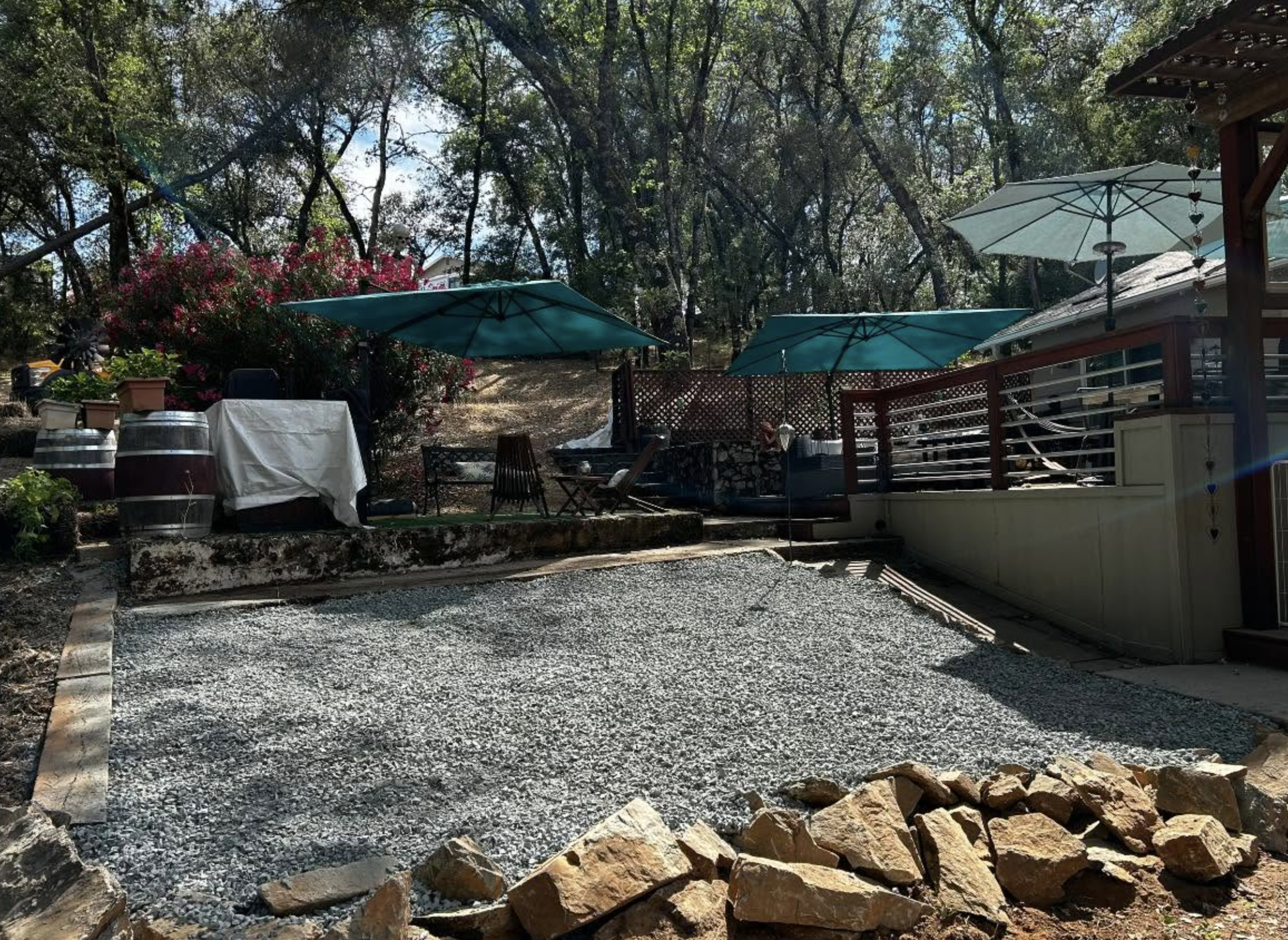 A backyard patio area with three large green umbrellas providing shade, surrounded by a wooden deck, potted plants, and tables covered with white cloth. The yard is filled with trees and bushes, with some wine barrels and a gravel surface in the foreground.