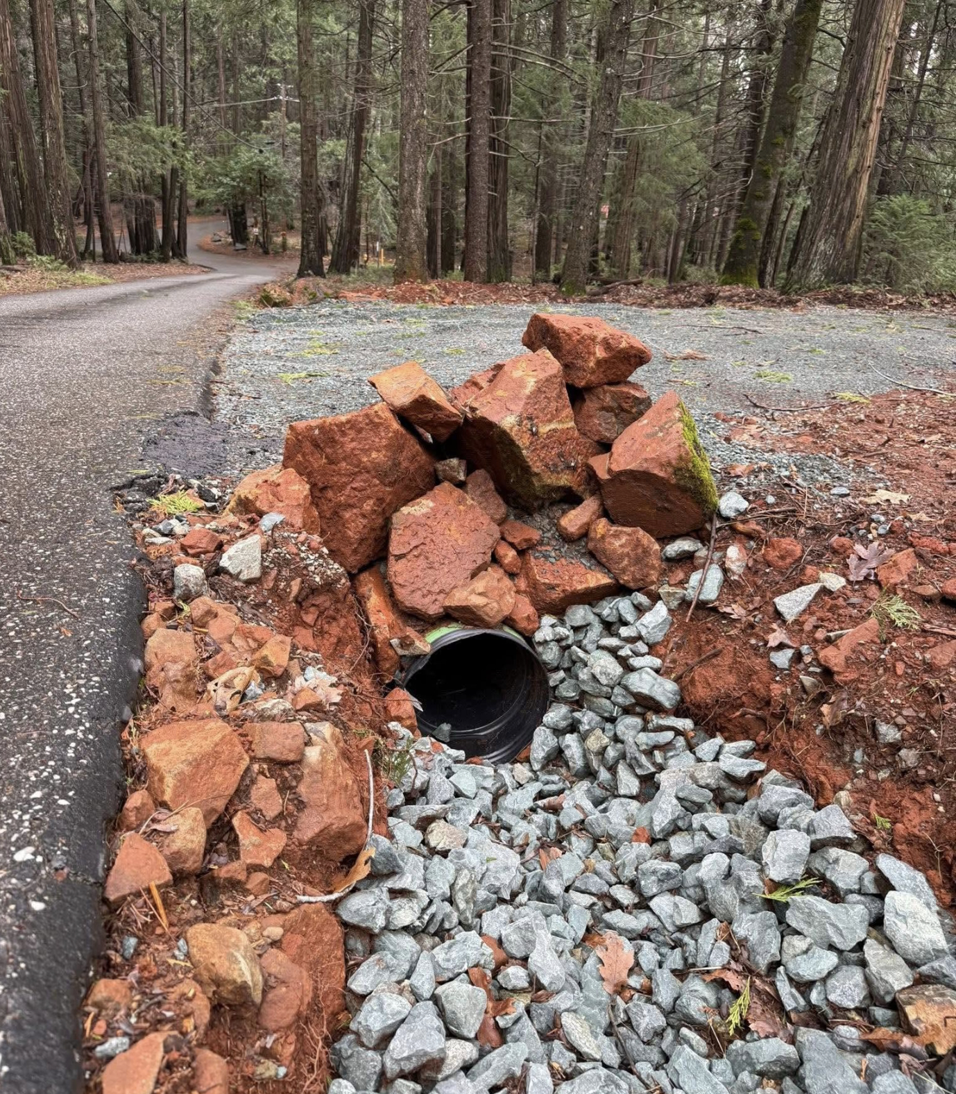 A partially constructed drainage pipe embedded in the ground along a woodland road, surrounded by rocks and dirt.