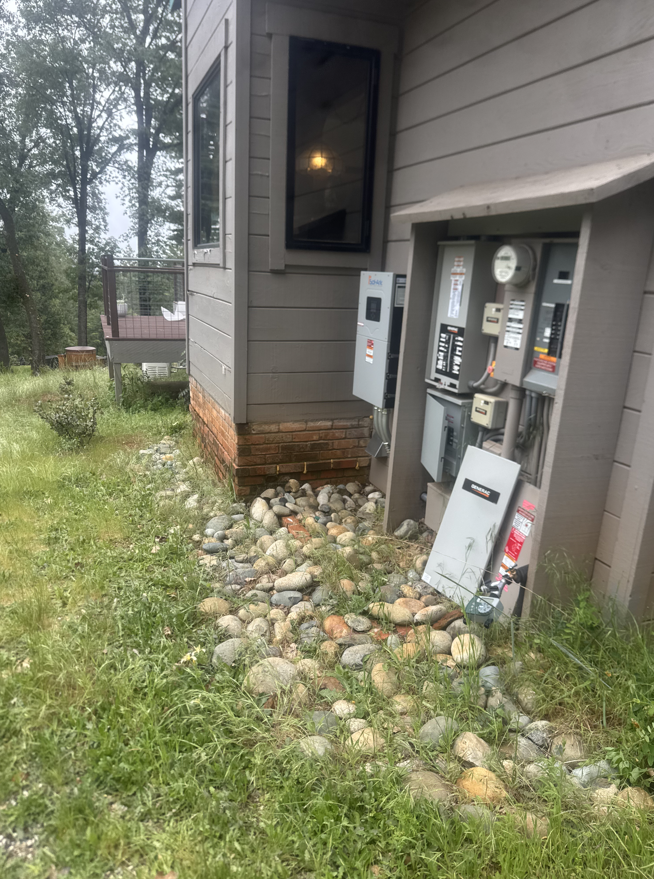 Side view of a house with electrical meters, a small balcony with a chair, and a yard with rocks and grass.