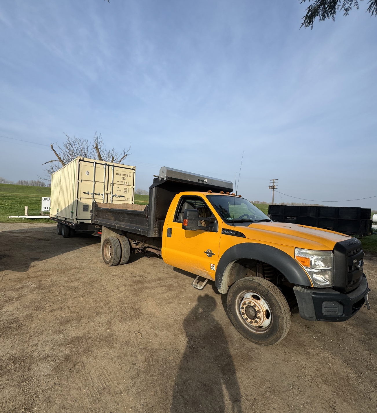 A yellow and black flatbed truck with a white shipping container on the back, parked outdoors on a dirt lot with grass and trees in the background under a blue sky.