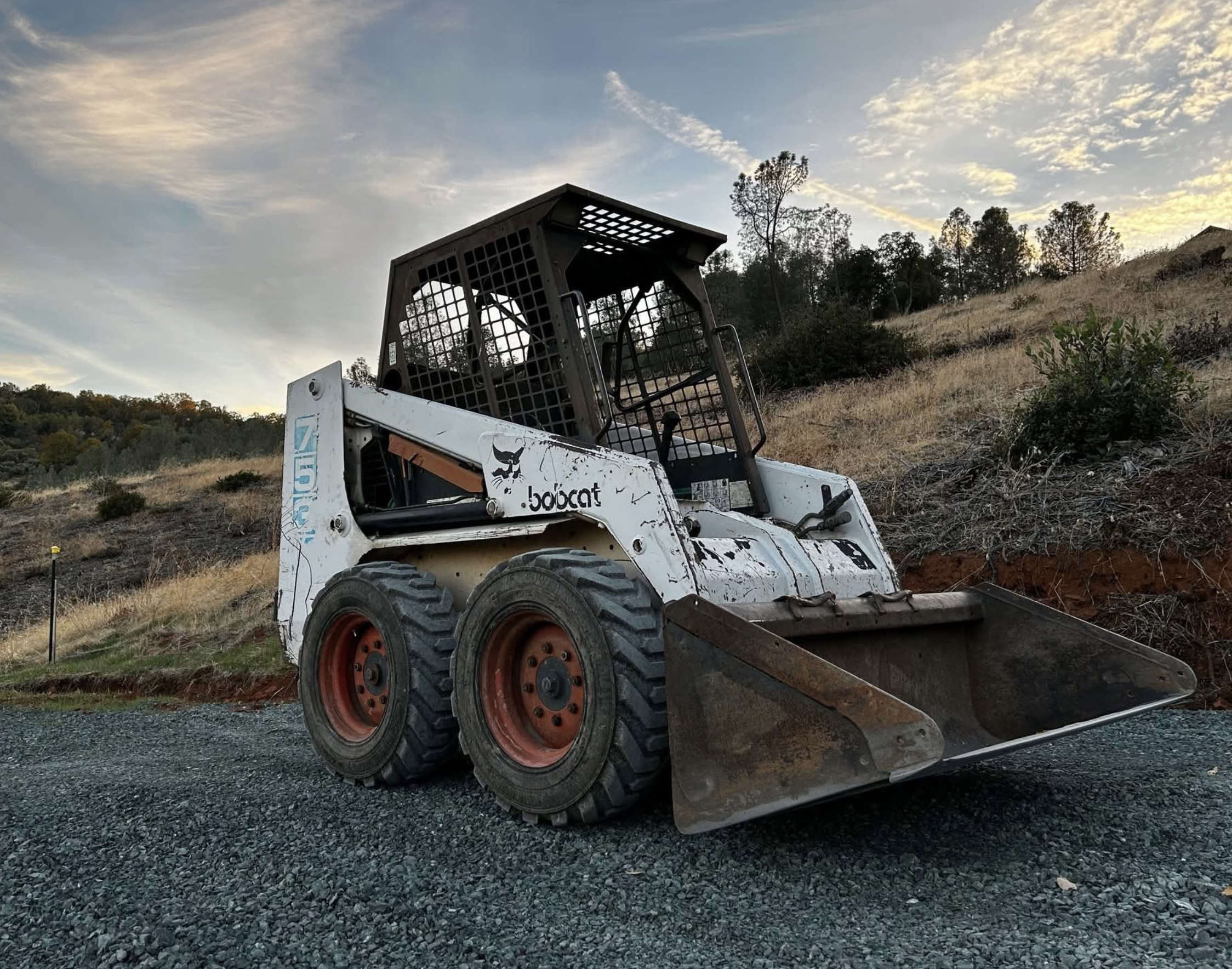A skid-steer loader on a gravel surface, with a hillside and trees in the background during what appears to be sunset, under a partly cloudy sky.