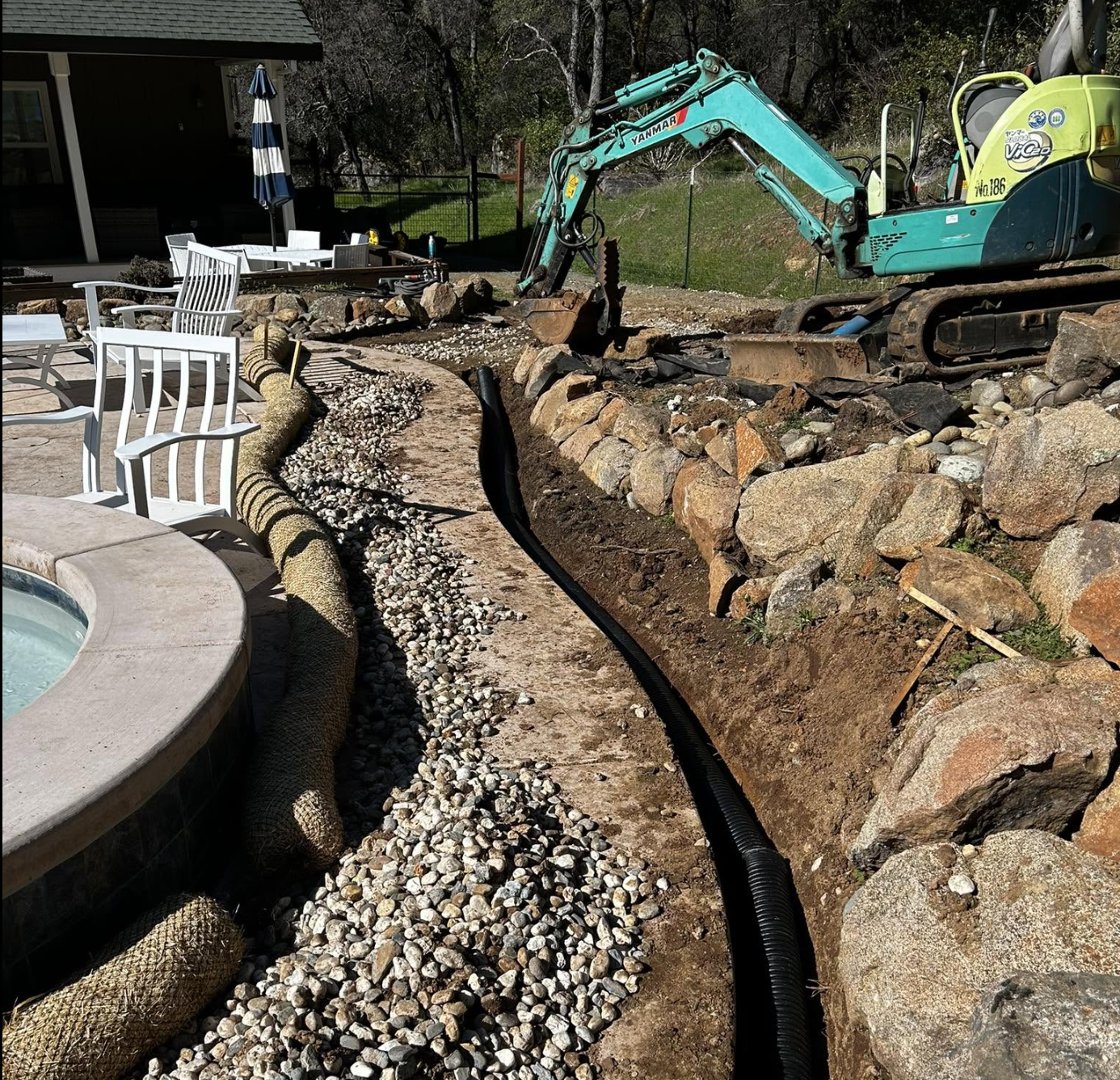 A backyard under construction with a small excavator, a stone border wall, and a partially installed landscape drainage system.