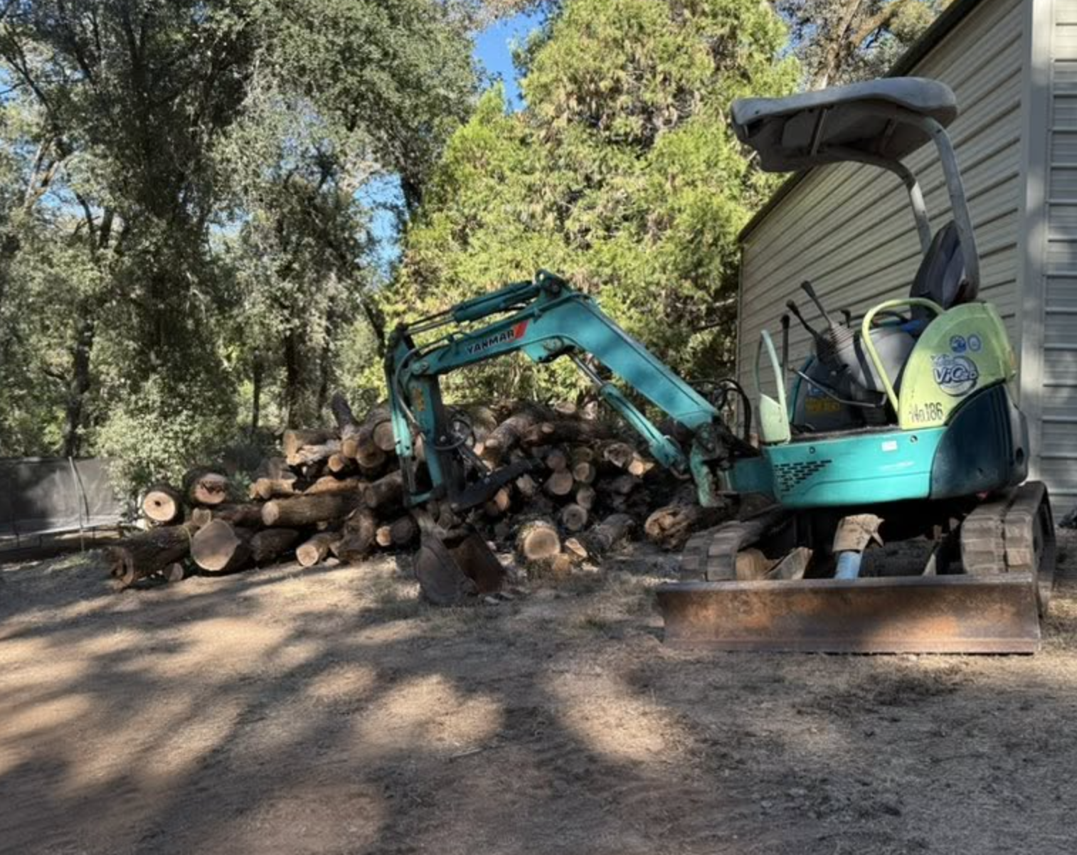 A small turquoise mini excavator sitting on a pile of cut logs near a beige building, with trees and blue sky in the background.