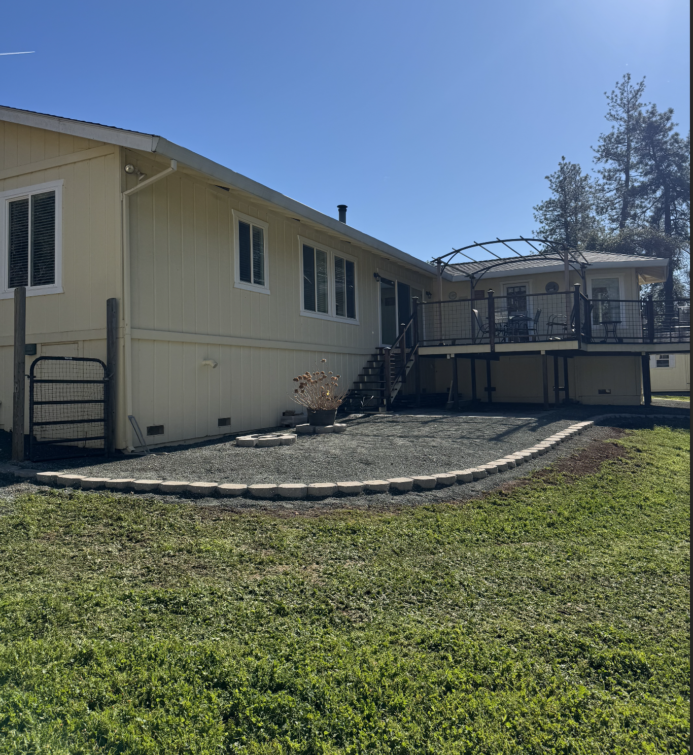 A beige house with a raised wooden deck, sliding glass door, and stairs leading down to a gravel patio area, surrounded by a grassy yard with a large tree in the background.
