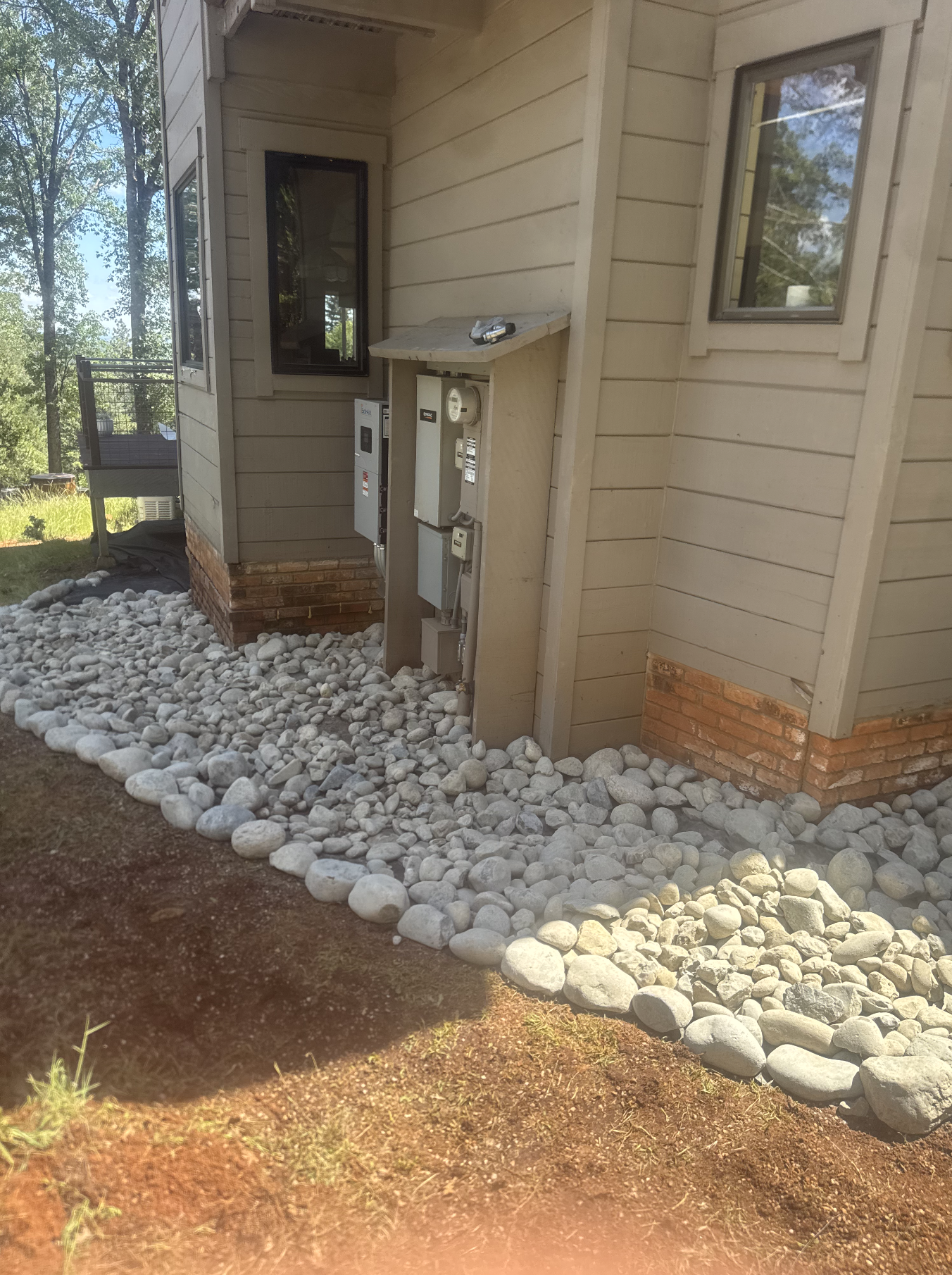 The exterior of a house with beige siding, two windows, and a brick foundation. In front, there is a layer of white rocks and some exposed soil.