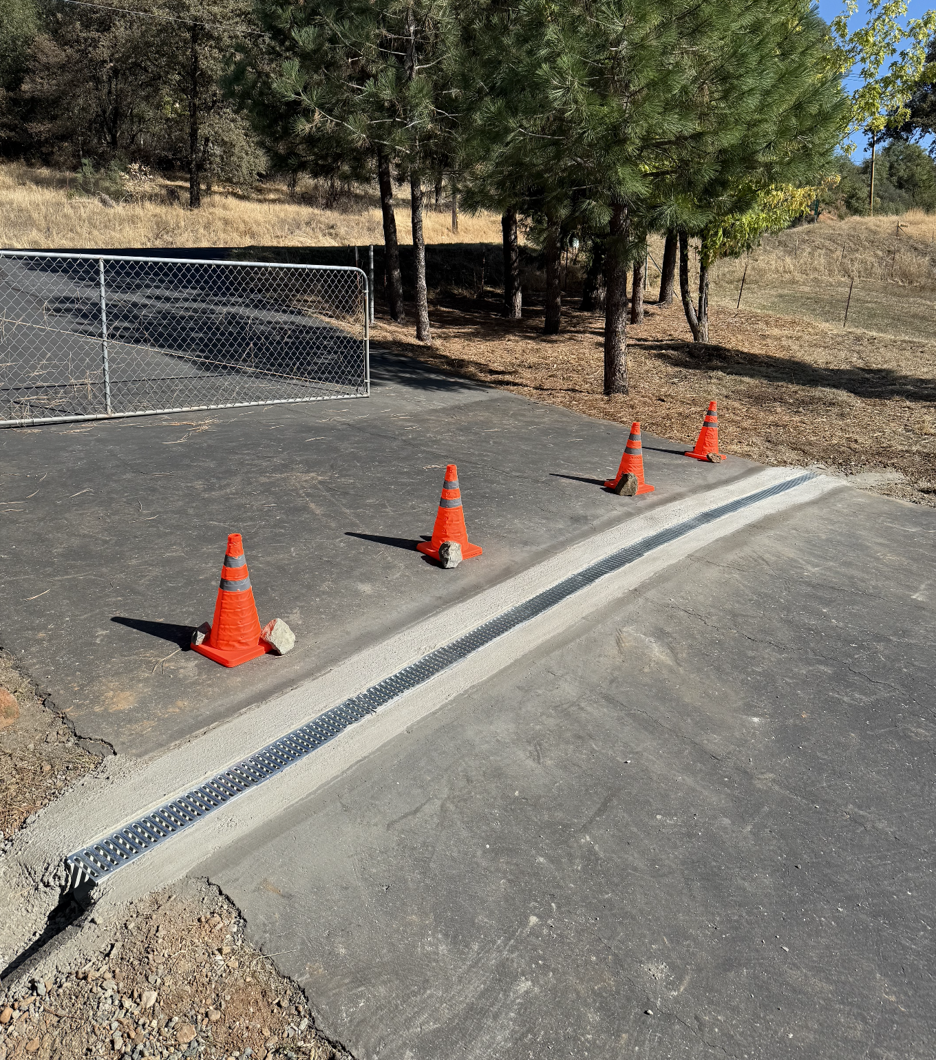 Four orange traffic cones with rocks on them blocking a section of freshly poured concrete in an outdoor area with trees and a chain-link fence.