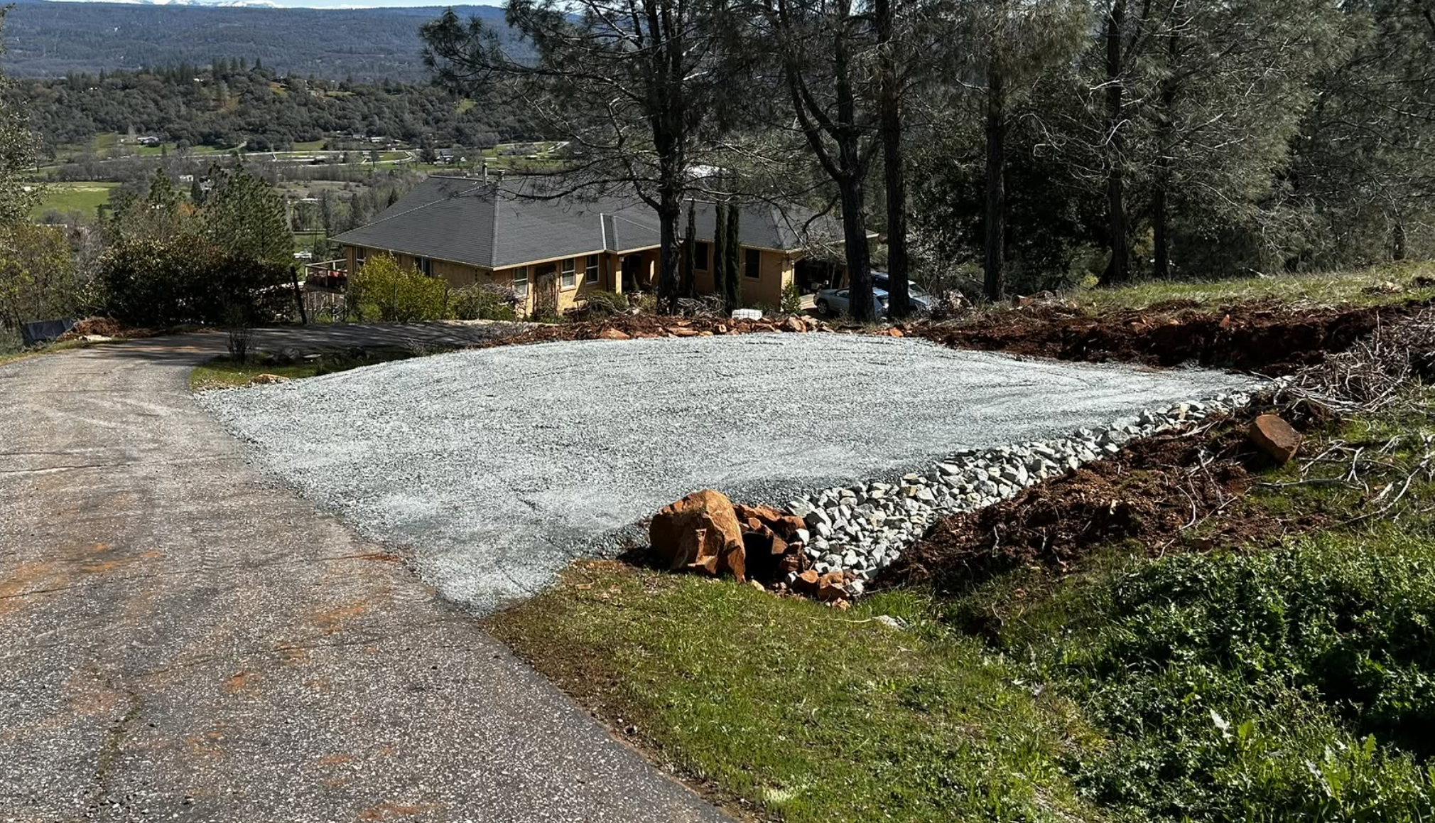 A gravel driveway on a hillside under construction with a yellow house and trees nearby, overlooking a valley with fields and hills in the distance.