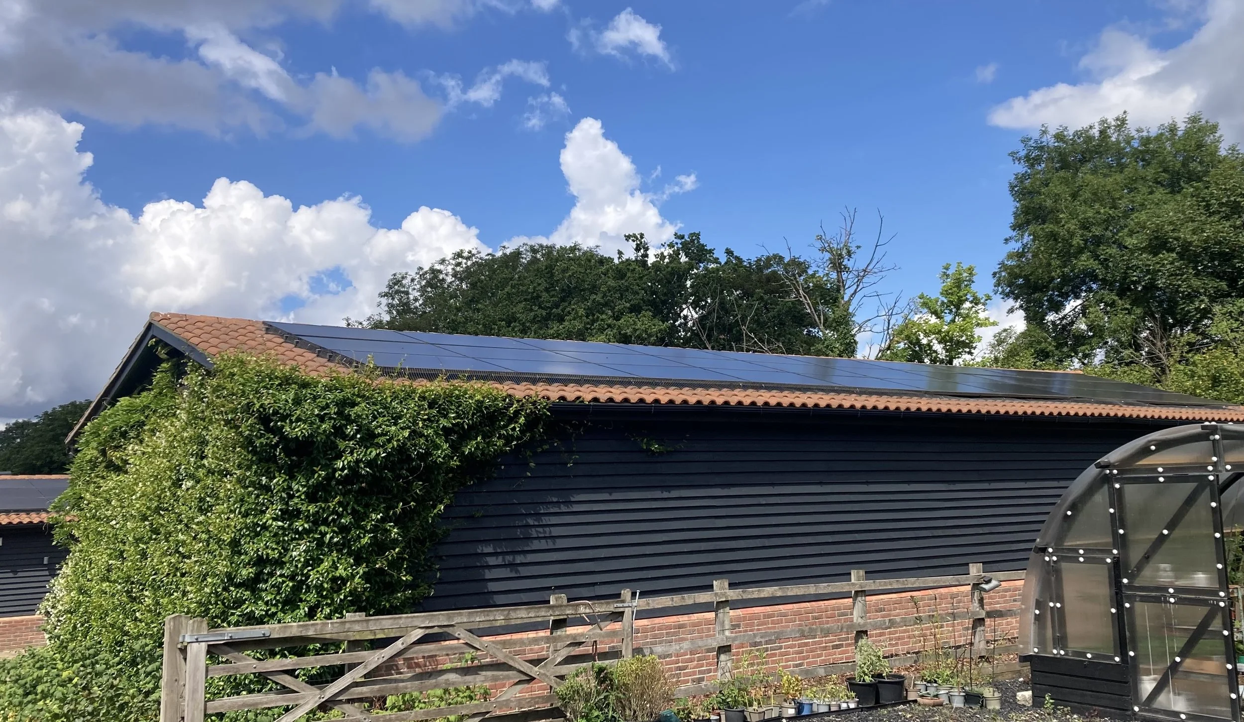 A house with a dark exterior and a red tiled roof, solar panels on the roof, surrounded by trees and greenery, with a blue sky and white clouds overhead.