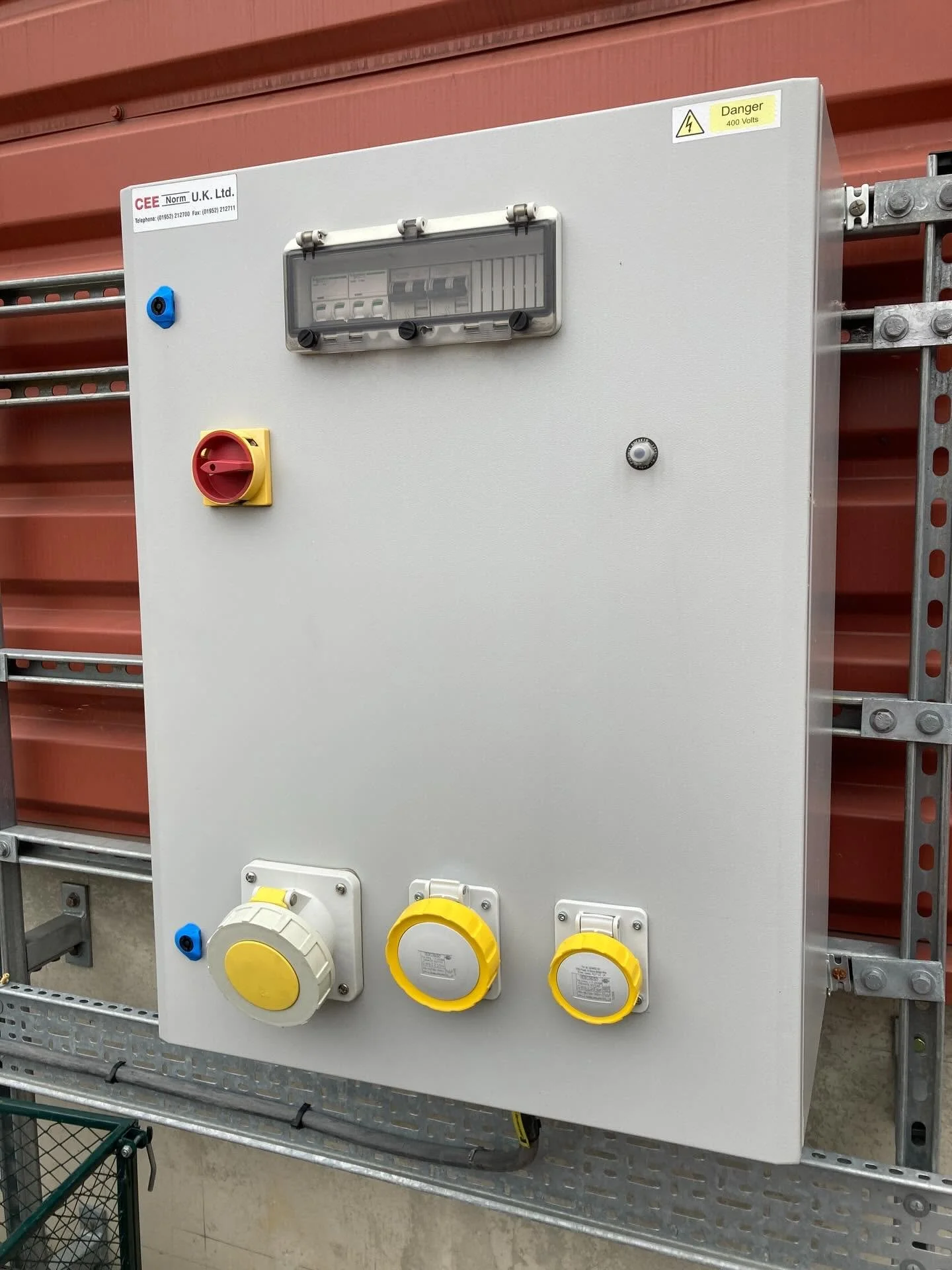 Electrical control panel with warning labels, switches, and power outlets, mounted on a metal rack.