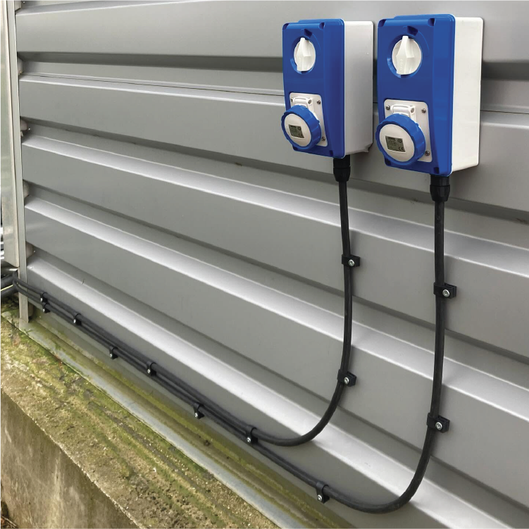 Two blue and white electric vehicle charging stations mounted on a gray metal exterior wall with black charging cables beneath.
