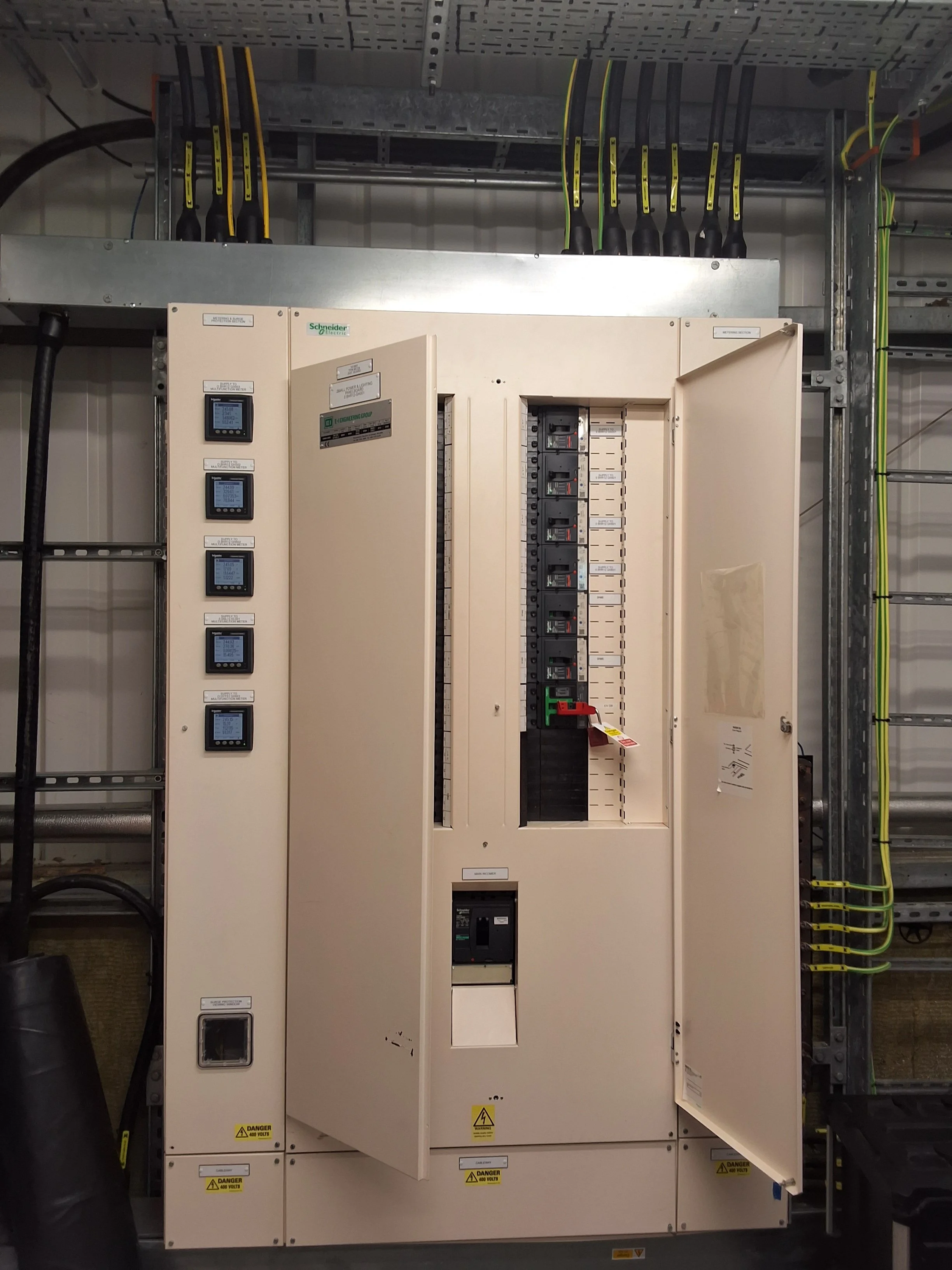 Electrical control panel with multiple circuit breakers, digital displays, and warning labels, mounted inside an industrial service enclosure, with electrical wiring and conduits visible around it.