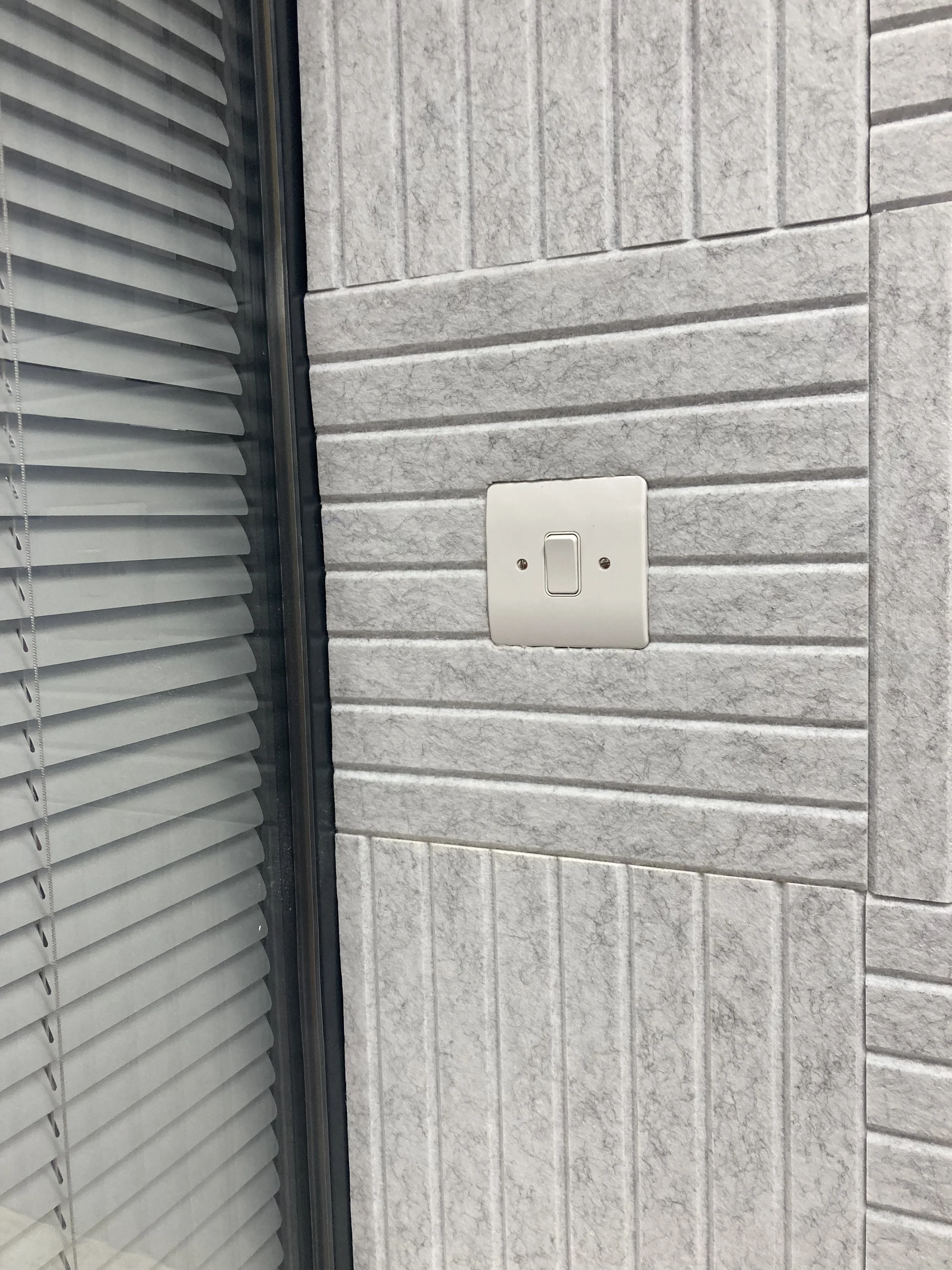 A section of a beige brick wall with a white light switch and a vertical window with closed horizontal blinds.