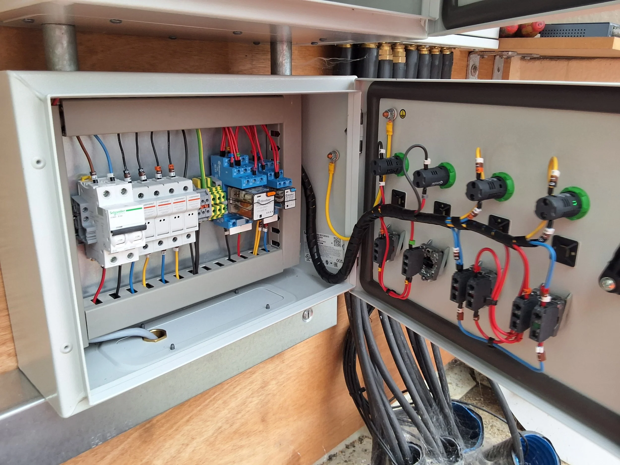 An electrical control panel with various wiring, circuit breakers, and connectors mounted inside a gray metal enclosure, with a neighboring panel featuring black and green connectors and cables.