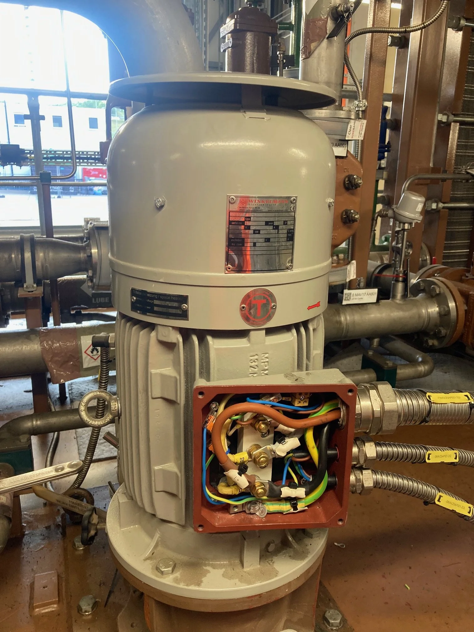 An industrial machine with an open panel, revealing internal wiring and components, situated in a manufacturing or mechanical room with pipes and metal structures.
