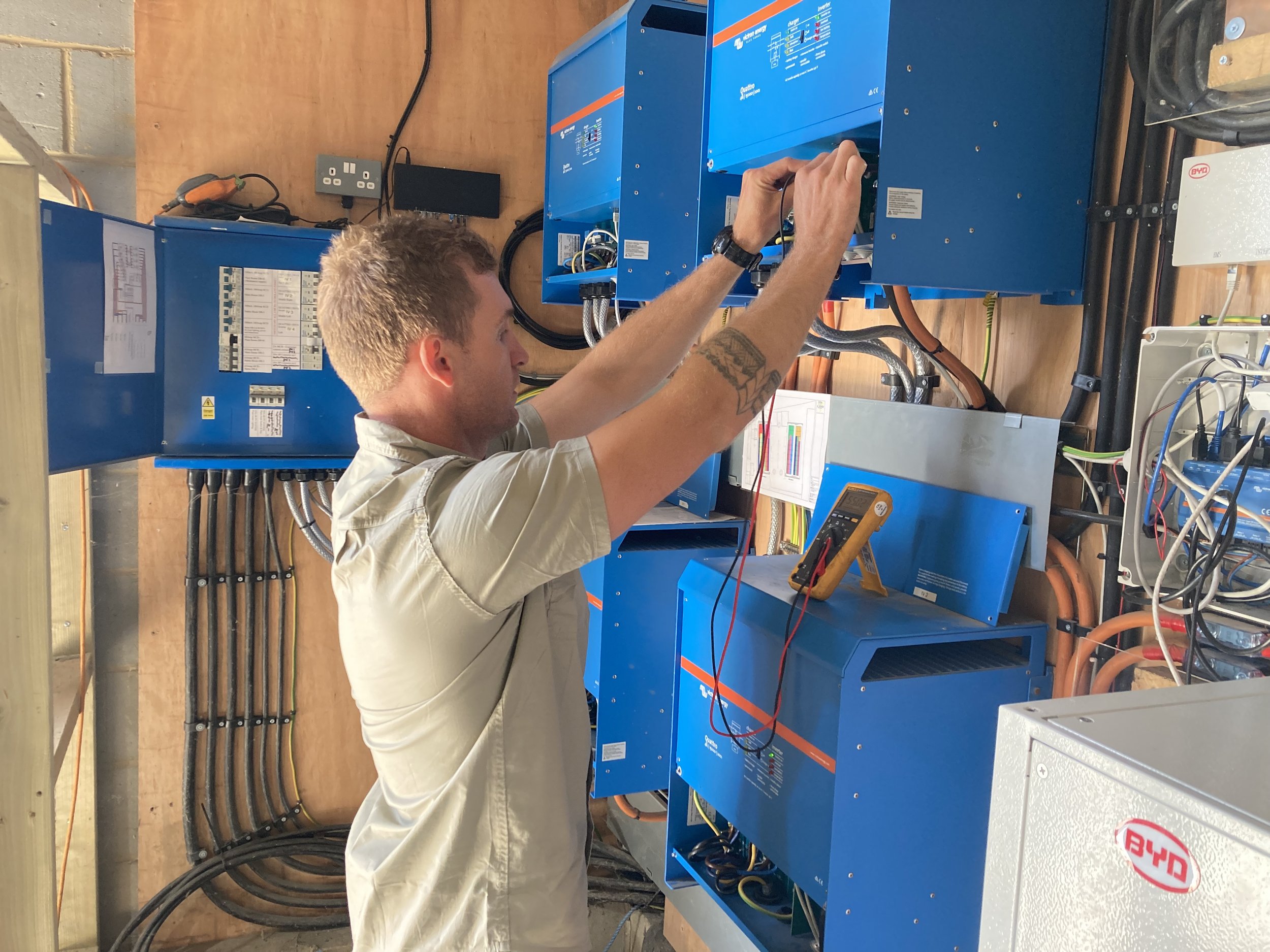 A technician repairing or inspecting electrical equipment in a utility room or electrical panel. He is working on blue electrical devices with wires and tools nearby.