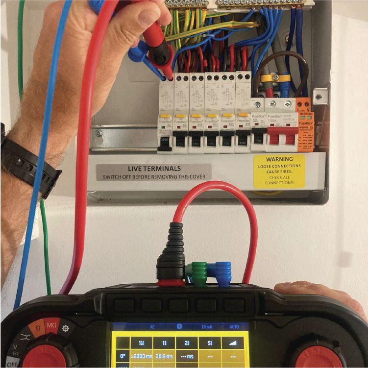 A person testing an electrical panel with a multimeter. The panel has circuit breakers and various wires connected.
