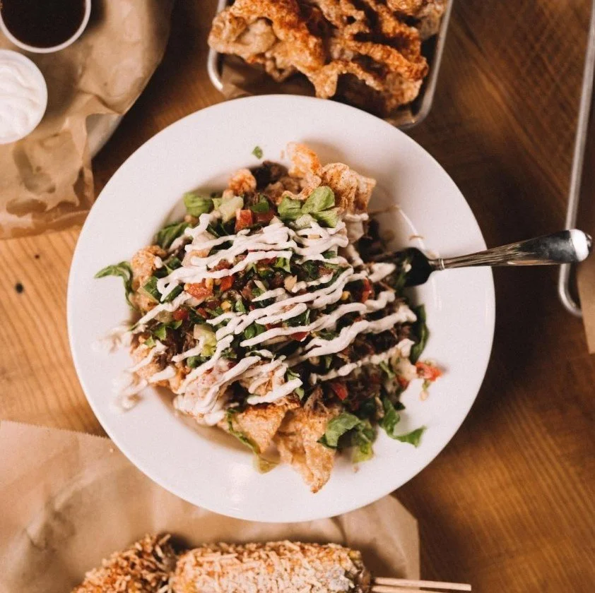 A white plate with chopped vegetables, lettuce, and drizzled with white sauce, on a wooden table. Pork Rind pieces are in a basket at the top right corner.