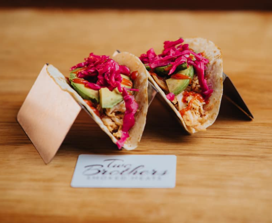 Two tacos filled with shredded chicken, avocado slices, and pink pickled vegetables, served on a wooden surface.