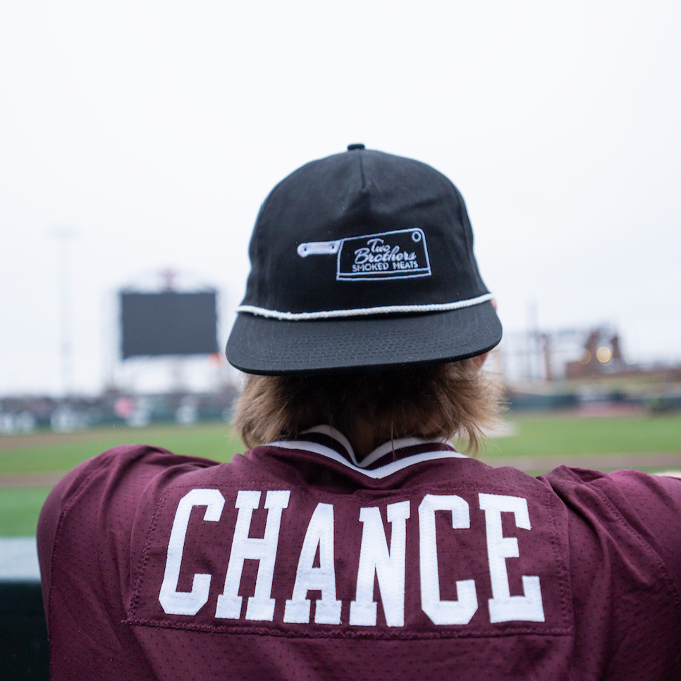Person wearing a maroon sports jersey with the word 'CHANCE' on the back, and a black cap, sitting in a sports stadium and looking at the field.