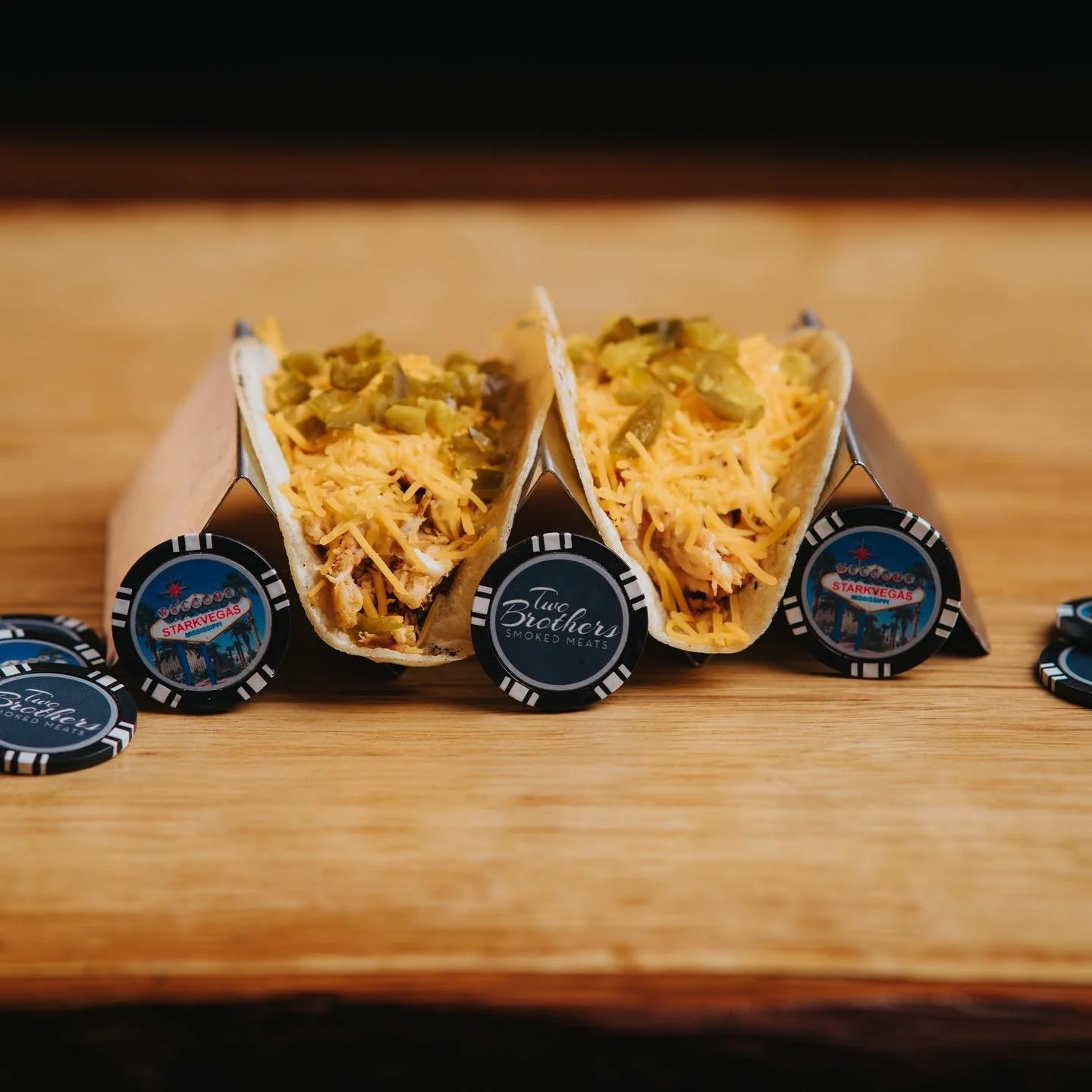 Two tacos filled with shredded cheese and chopped green chiles, placed on a wooden surface with poker chips labeled 'Two Brothers Smoked Meats' and 'StarkVegas' surrounding them.