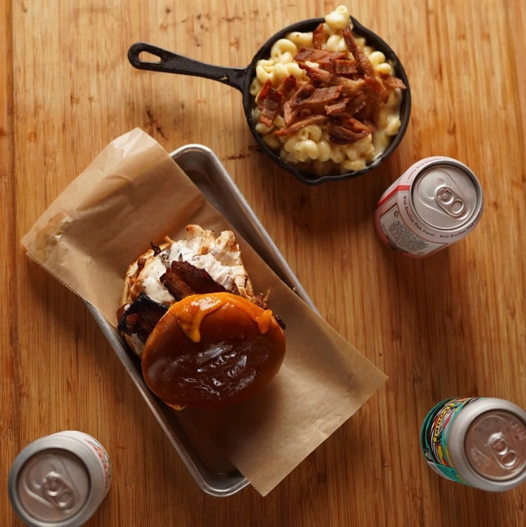 A white bbq chicken sandwich on a metal tray with parchment paper, a small cast-iron skillet of macaroni and cheese with bacon, and three cans of soda on a wooden table.
