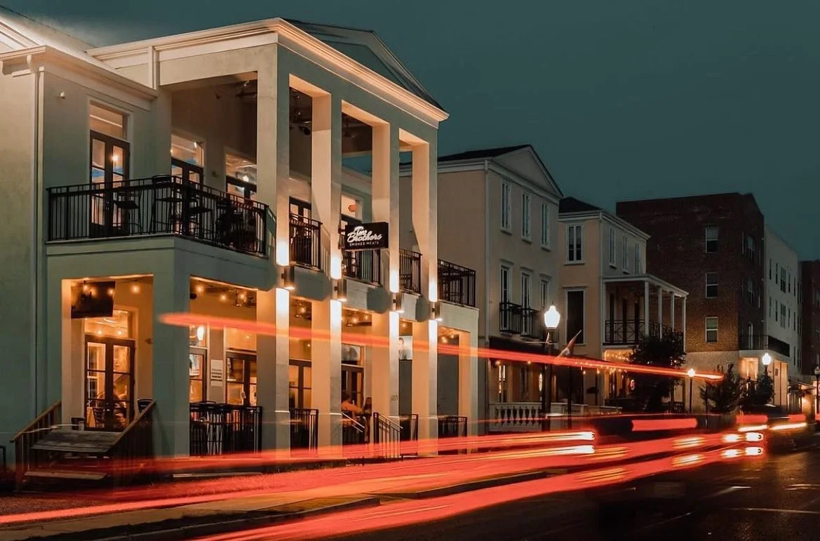Street view at night with a modern, light green, two-story building housing a restaurant called 'Two Brothers' with outdoor balconies illuminated by warm lighting. Light trails from passing vehicles streak across the image.