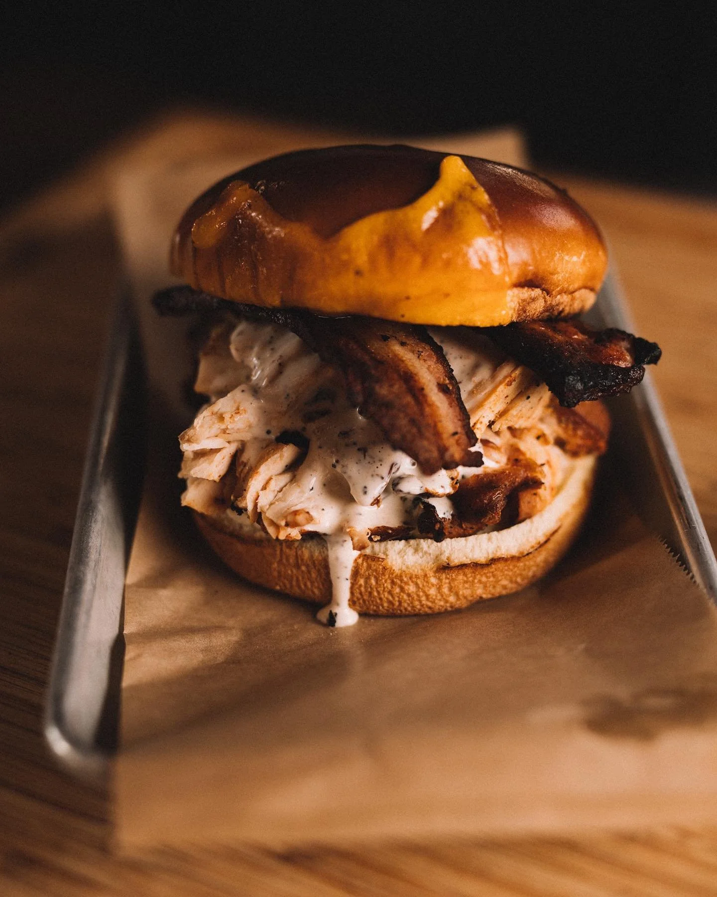 A gourmet sandwich with grilled chicken, crispy bacon, and a toasted bun, served on a metal tray with parchment paper.