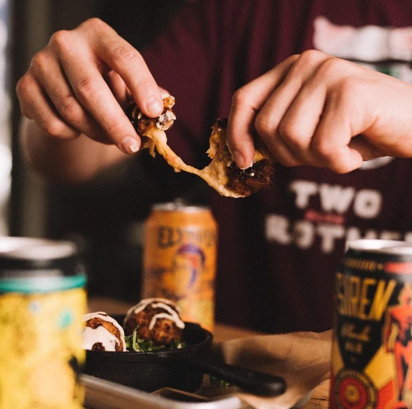 Person tearing apart a piece of fried pimento cheese, with cans of beer and a small plate of  fried pimento cheese with sauce in the foreground.