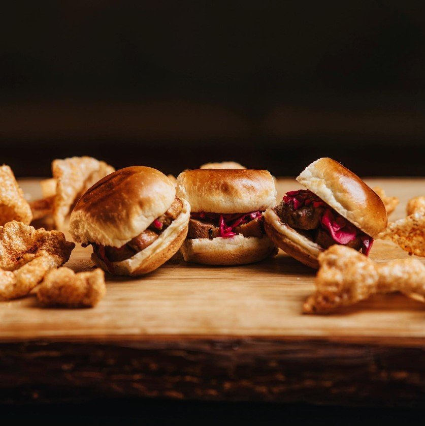 Three mini sandwiches with pork tenderloin, pickled red onions, on a wooden serving board, surrounded by chicken nuggets.