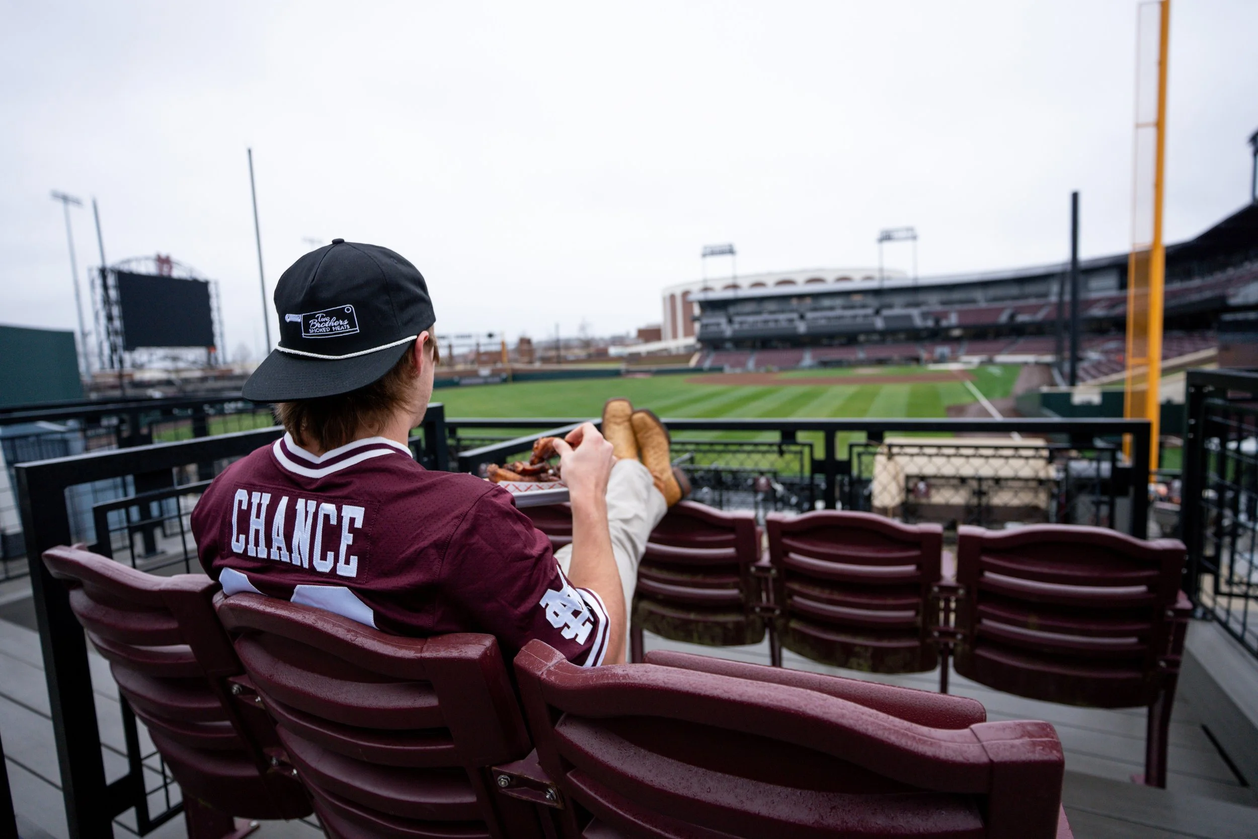 A person sitting in stadium seats at an empty baseball stadium, wearing a maroon and white jersey with 'CHANCE' on the back, holding a tray chicken wing, with the field and stadium infrastructure visible in the background.
