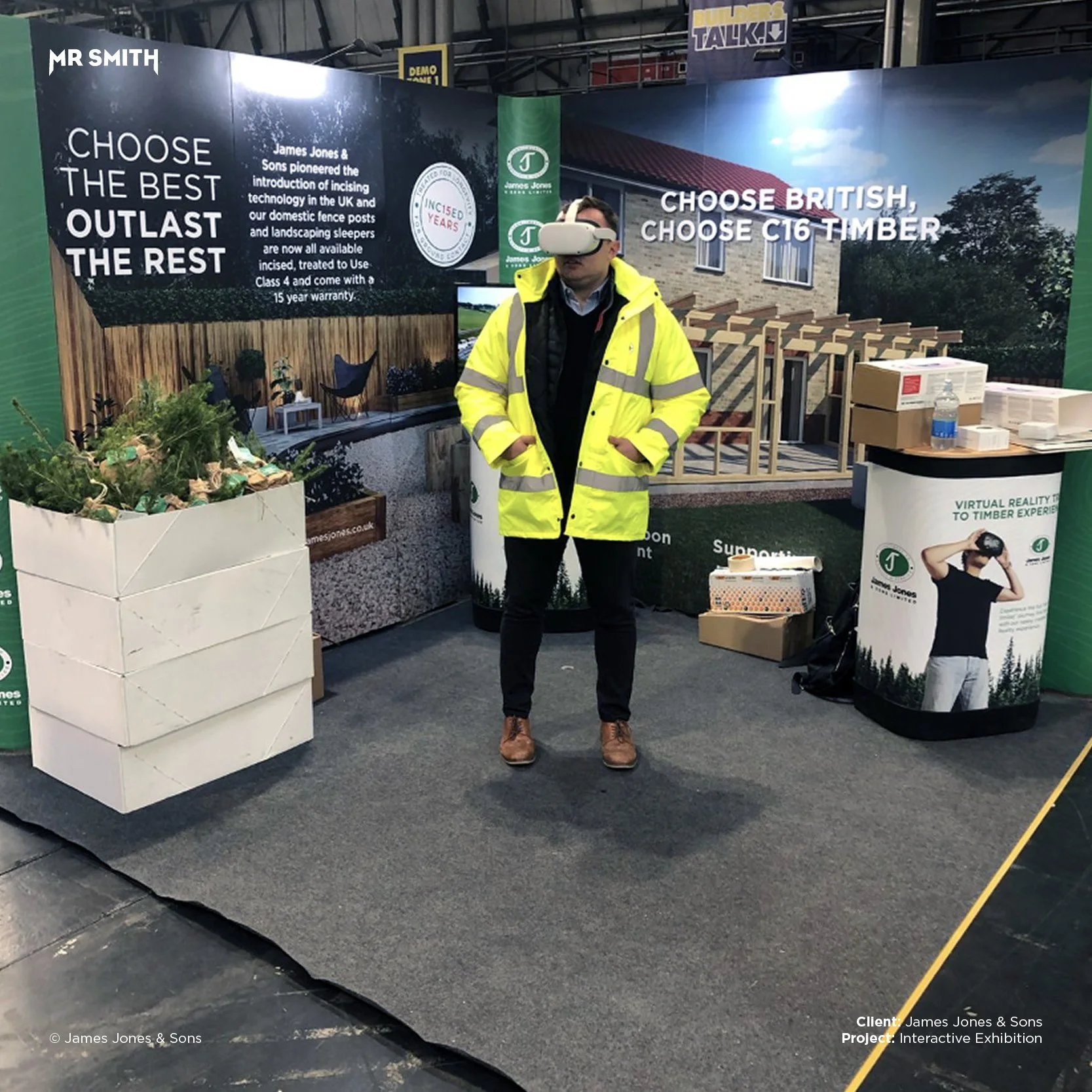 A man wearing a yellow safety jacket and a virtual reality headset standing at an exhibition booth promoting timber products. The booth features a large graphic of a house and various greenery, with signs that say 'Choose the best outlast the rest' a