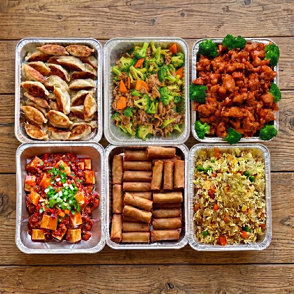 Six aluminum containers with various Asian dishes arranged on a wooden table. Top row has potstickers, stir-fried broccoli and beef, and sweet and sour chicken. Bottom row has General Tso's tofu, spring rolls, and vegetable pasta salad.